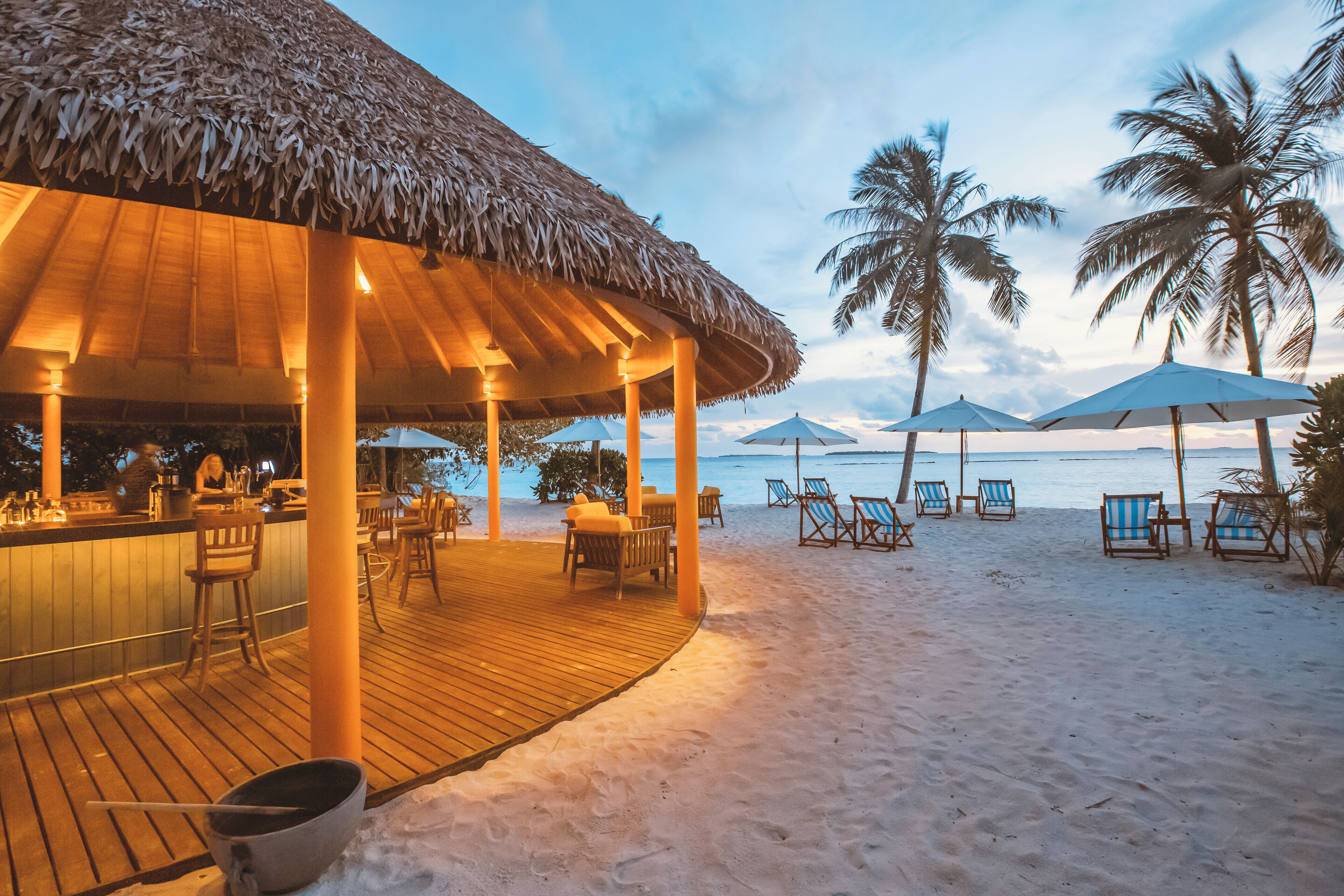 Eine Strandbar mit Holzterrasse und Strohdach, umgeben von Liegestühlen und Sonnenschirmen, im Hintergrund Palmen und das Meer.