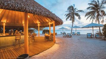 Eine Strandbar mit Holzterrasse und Strohdach, umgeben von Liegestühlen und Sonnenschirmen, im Hintergrund Palmen und das Meer.