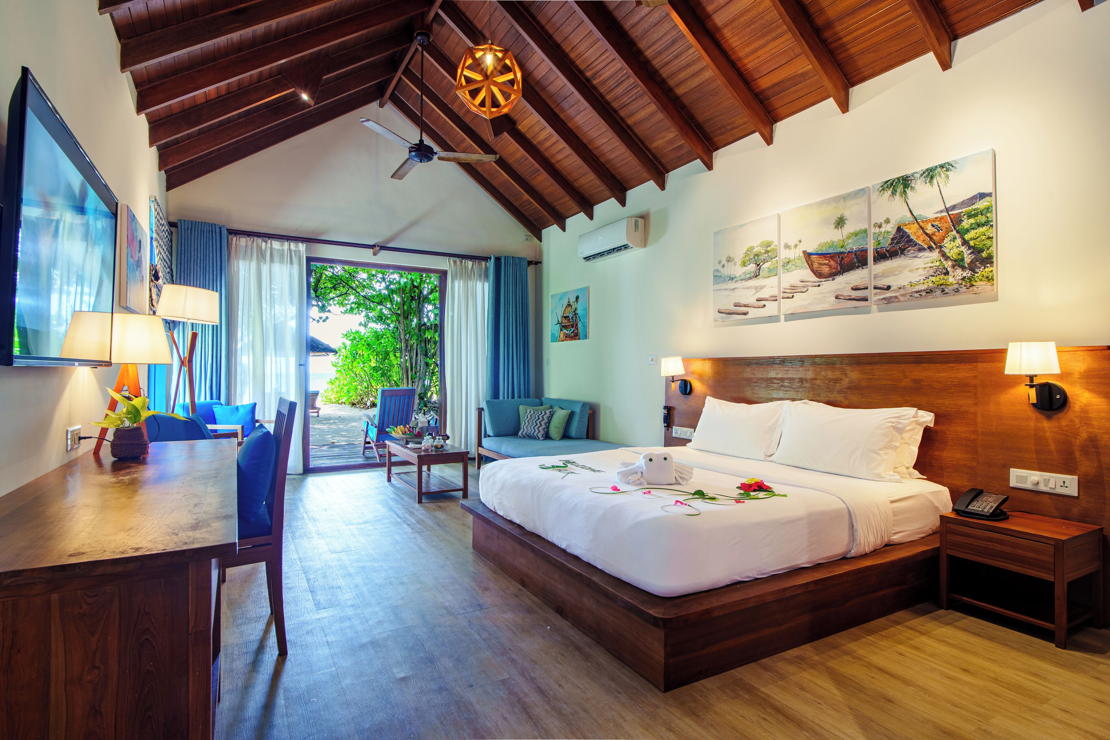 A hotel room with a wooden ceiling, a large bed, a desk with a lamp, a chair, a sofa, and a view of greenery outside.