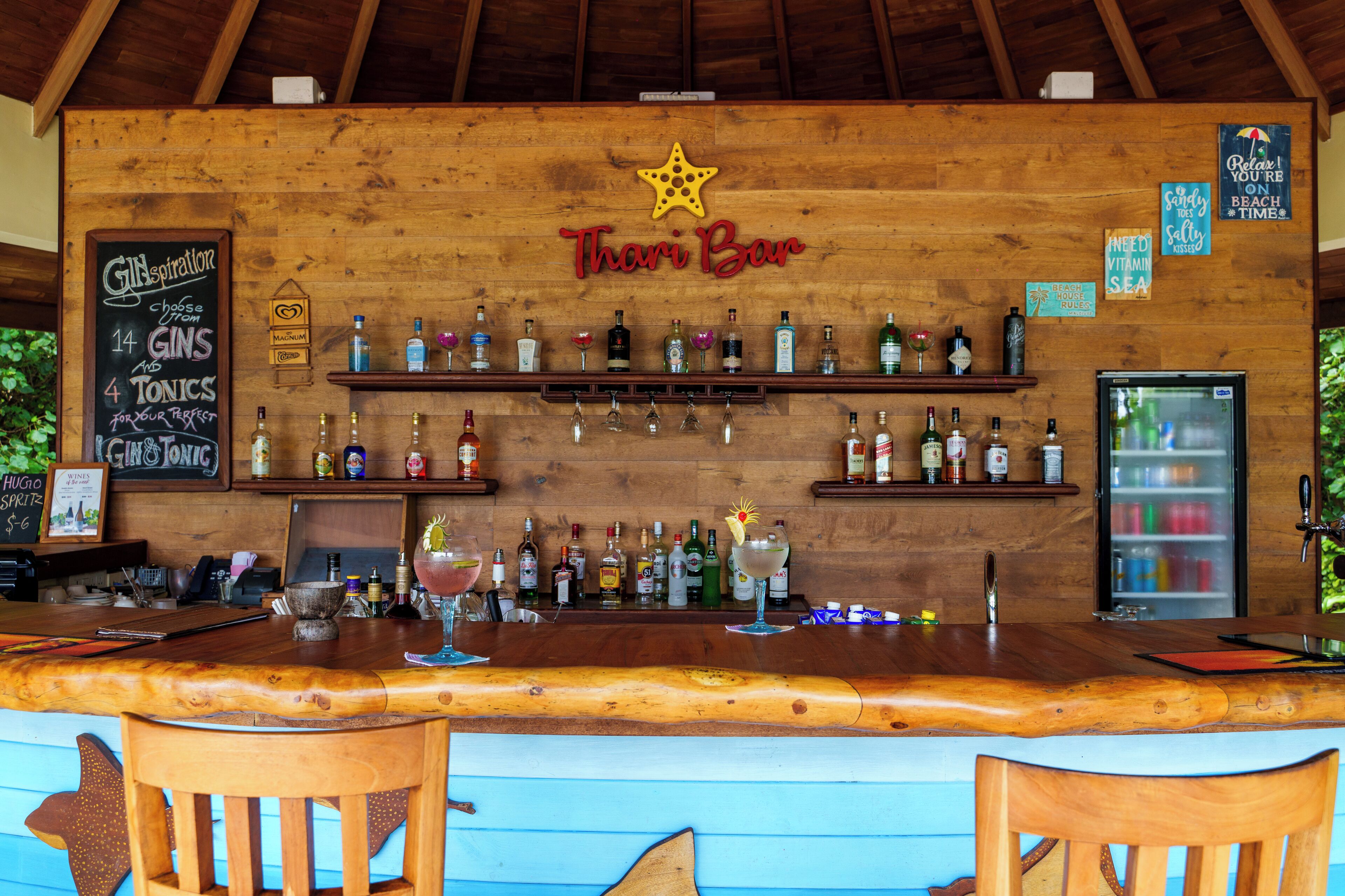Una zona de bar con mostrador de madera, estantes con botellas y un letrero de neón que dice "Thari Bar".