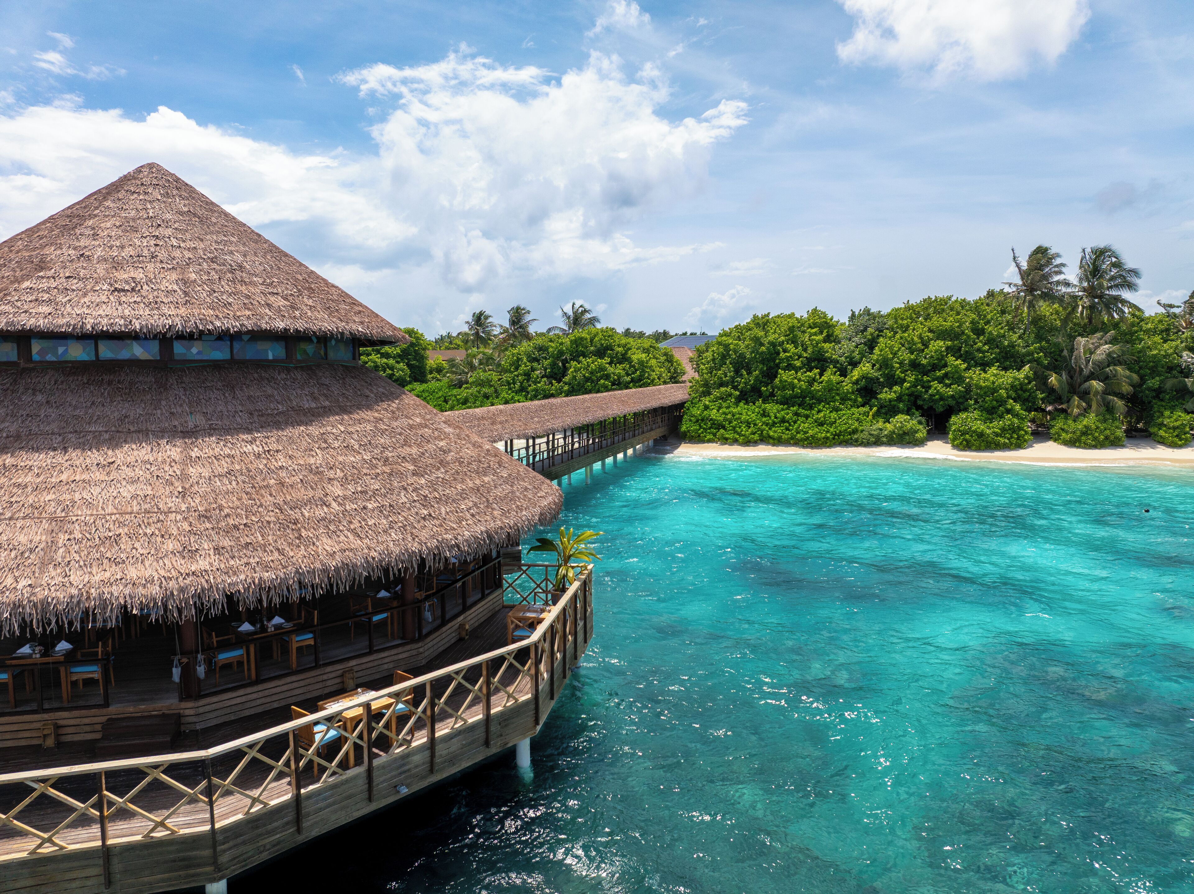 A thatched-roof overwater bungalow with a wooden deck and a view of turquoise waters and a sandy beach.