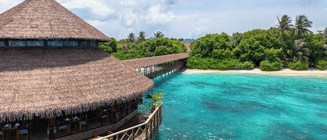 A thatched-roof overwater bungalow with a wooden deck and a view of turquoise waters and a sandy beach.