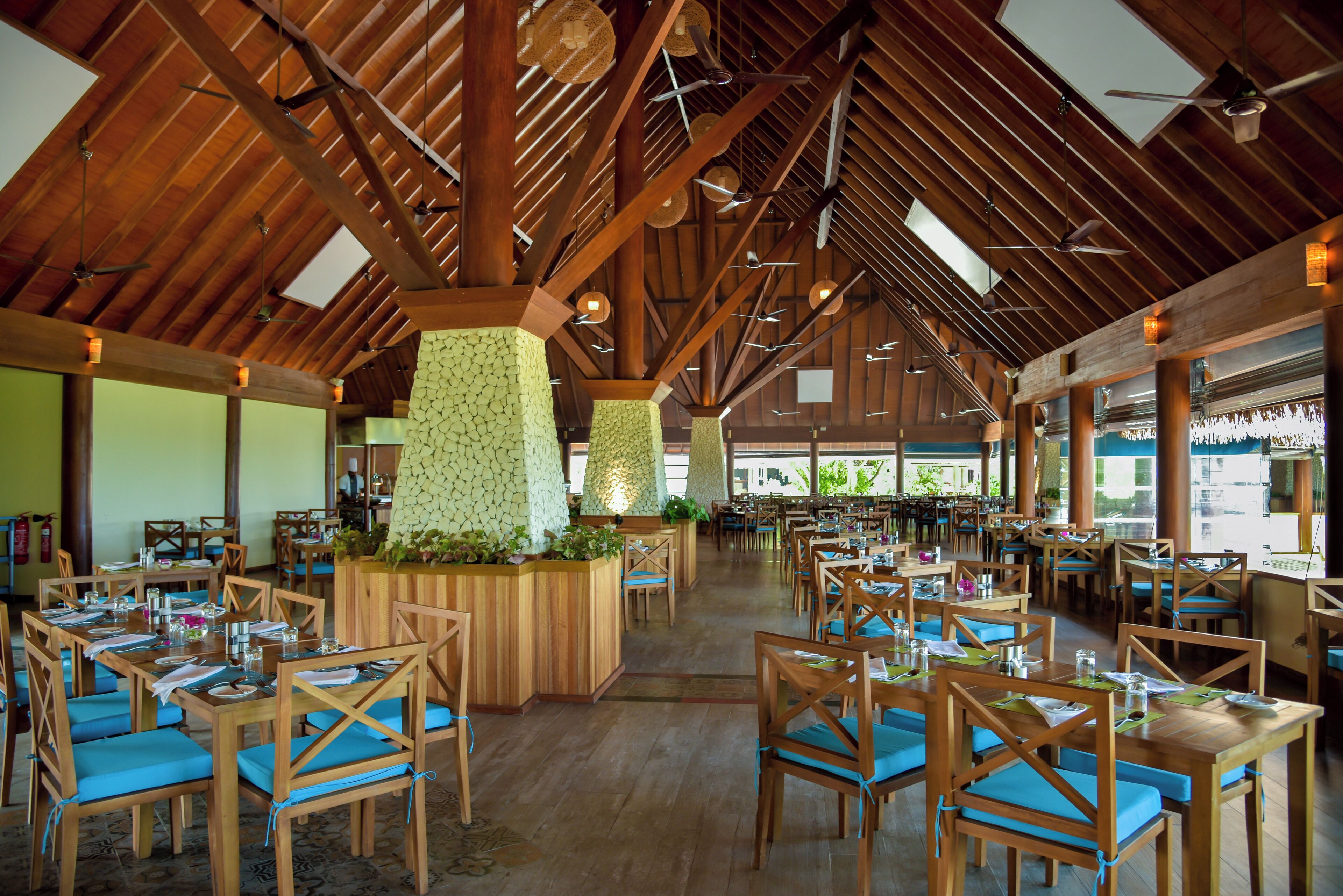 Un restaurante espacioso con mesas de madera y sillas con cojines azules, un techo alto de madera con tragaluces y una pared central decorada con vegetación.