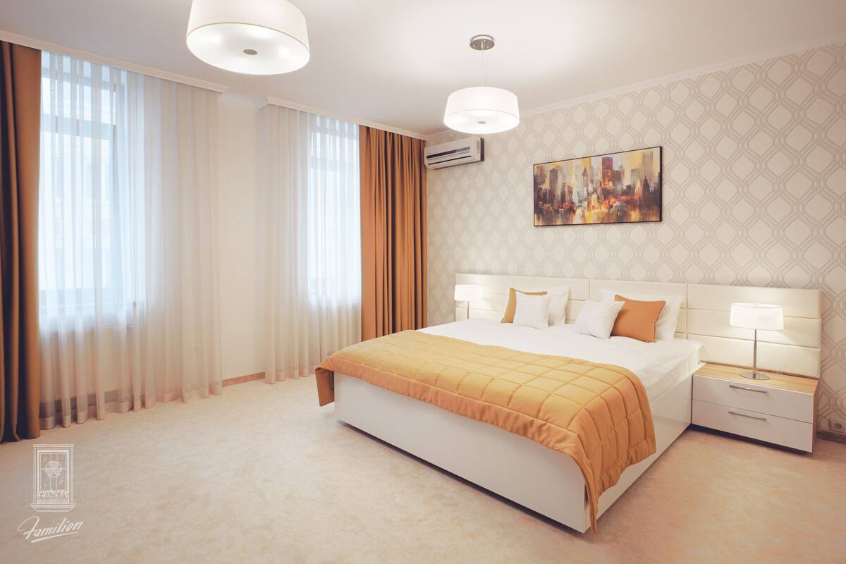 A modern hotel bedroom with a large bed, bedside lamps, a cityscape painting, and a window with sheer curtains.
