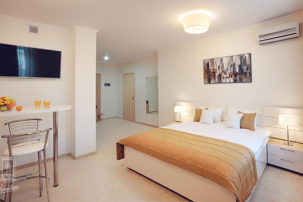 A hotel room with a bed, bedside tables, a TV, a desk with fruit and glasses, and a painting on the wall.