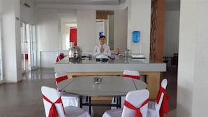 A modern hotel dining area with a central island, white chairs with red sashes, and a person standing behind the island. - Bangka City Hotel (Pangkalpinang)