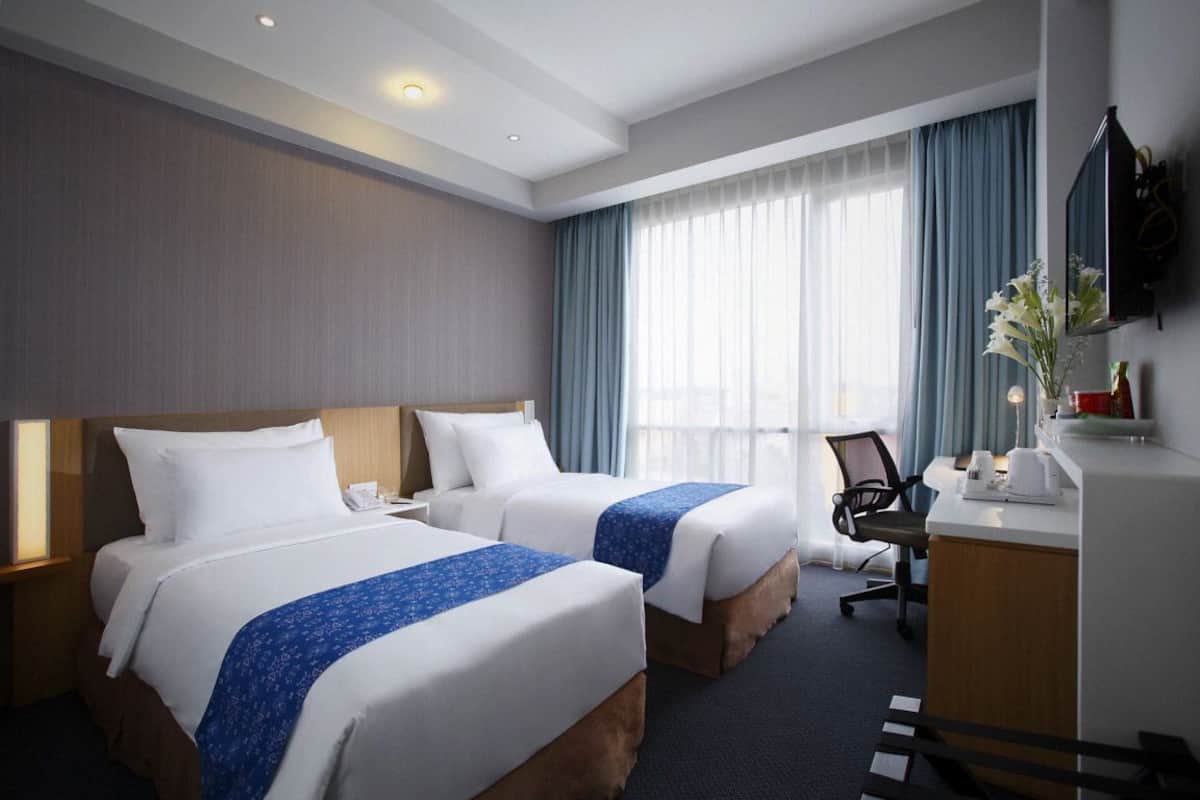 A hotel room with two beds, a desk, a chair, a TV, and a window with curtains.