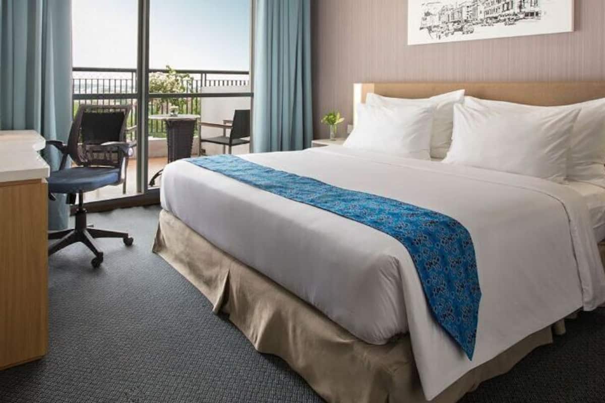 A hotel room with a bed, desk, chair, and a balcony with a view.
