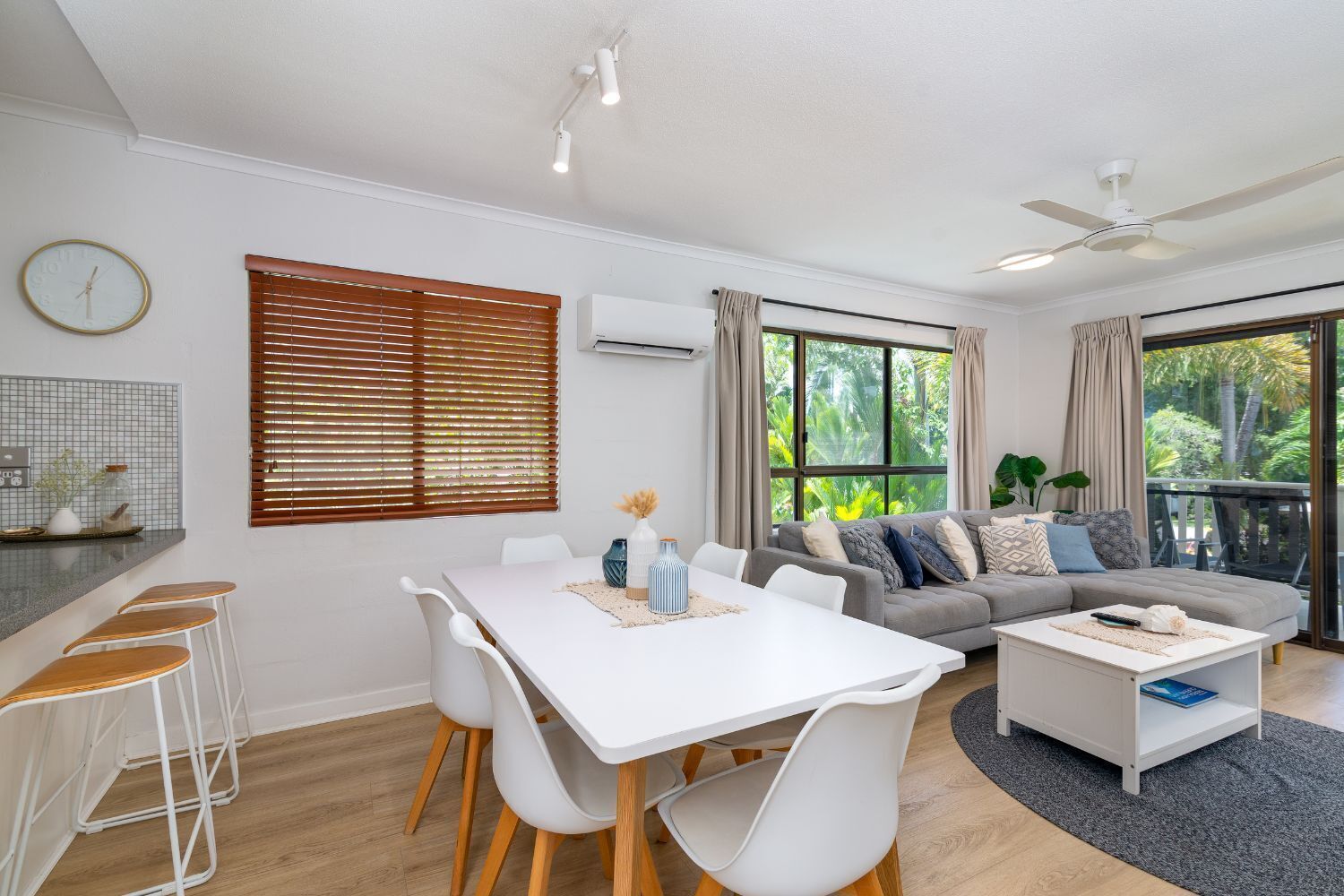 Een moderne woonkamer met een eettafel, witte stoelen, een grijze bank en een balkon met uitzicht op groen.