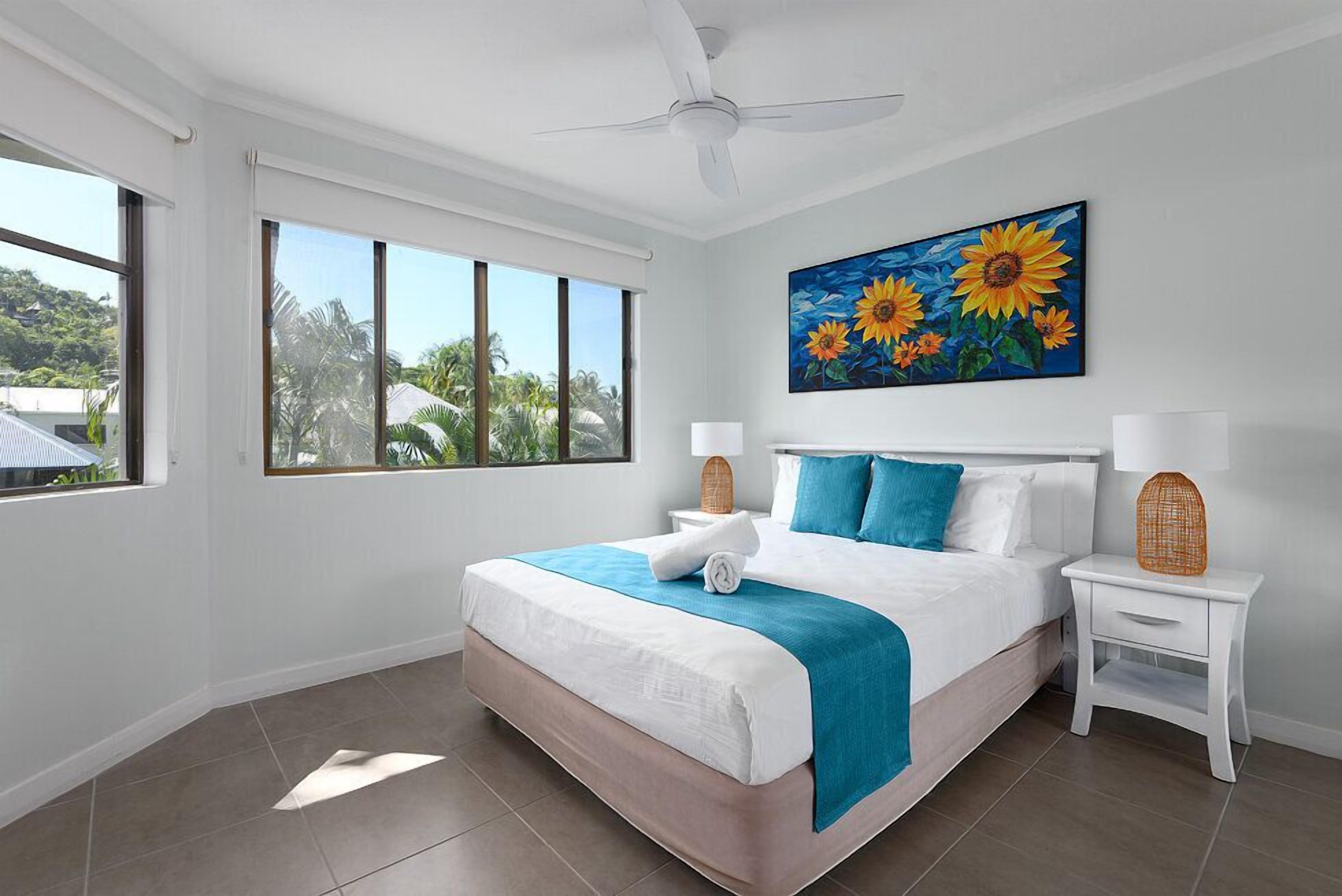 A hotel room with a bed, a ceiling fan, a painting of sunflowers, a window with a view of palm trees, and a nightstand with a lamp.
