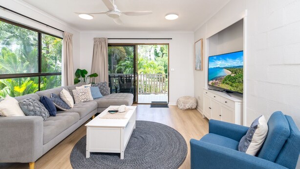 En moderne stue med sofa, sofabord og fjernsyn, samt en balkon med udsigt over grønne områder og en loftventilator.