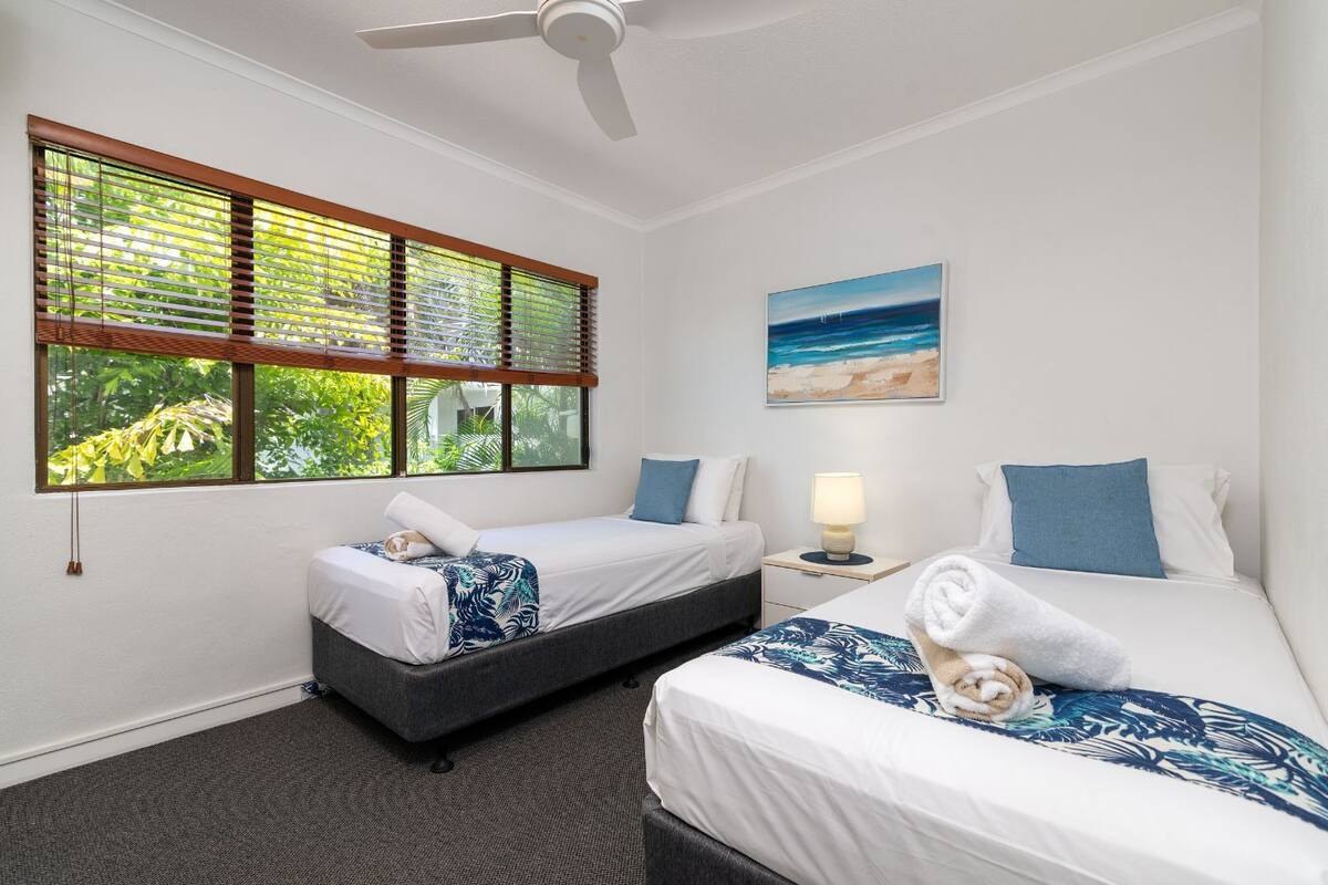 Ein Hotelzimmer mit zwei Betten, einem Deckenventilator, einem Fenster mit Jalousien, einer Nachttischlampe und einem Strandbild.