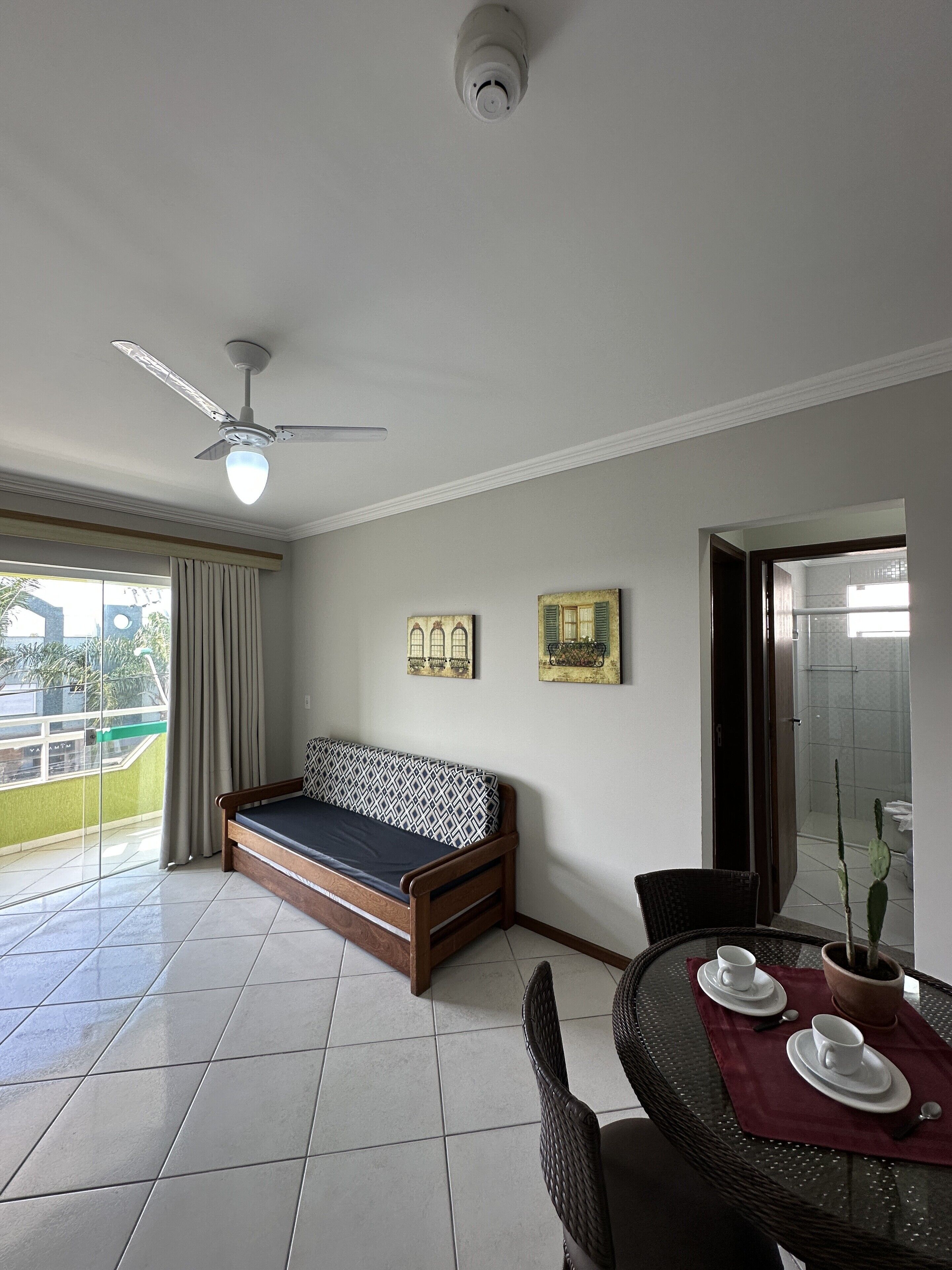 A room with a sofa, a dining table with cups and saucers, a ceiling fan, and a view of the outside.