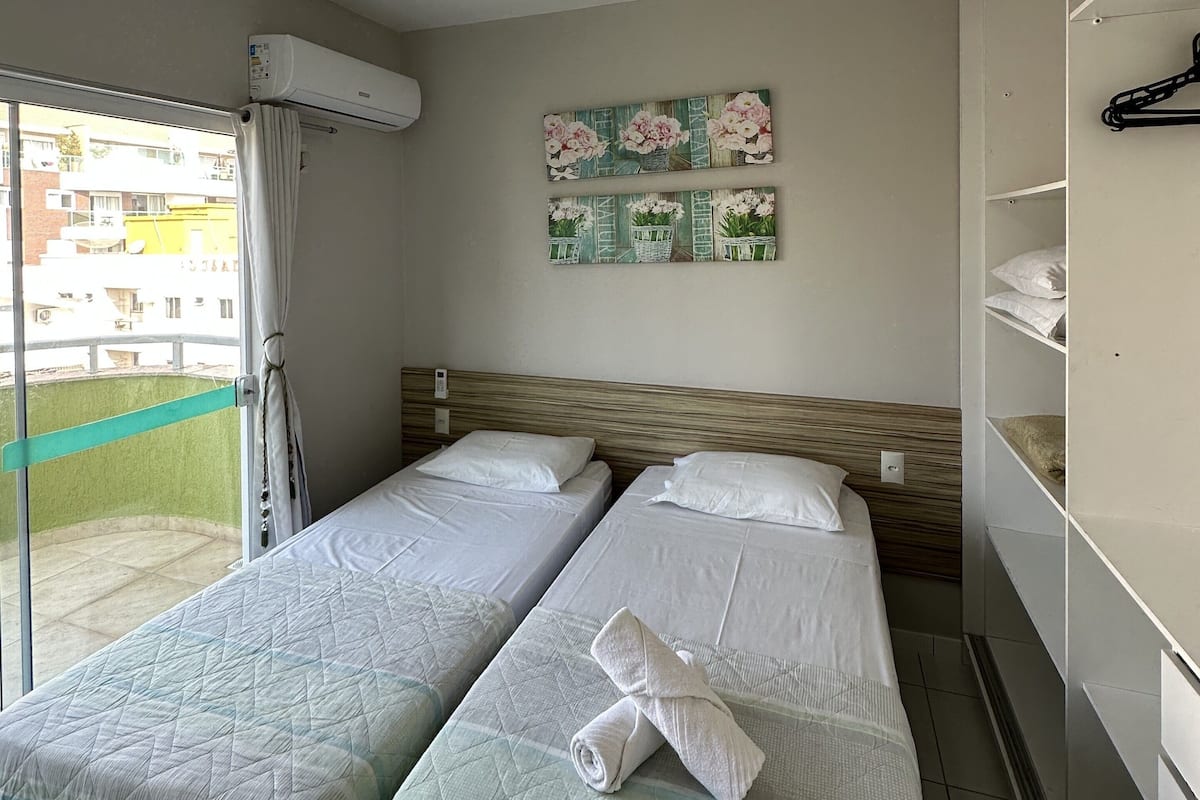 Hotellihuone, jossa on kaksi sänkyä, lasiliukuovi parvekkeelle, seinään asennettu ilmastointilaite ja hylly, jossa on siististi taiteltuja pyyhkeitä.