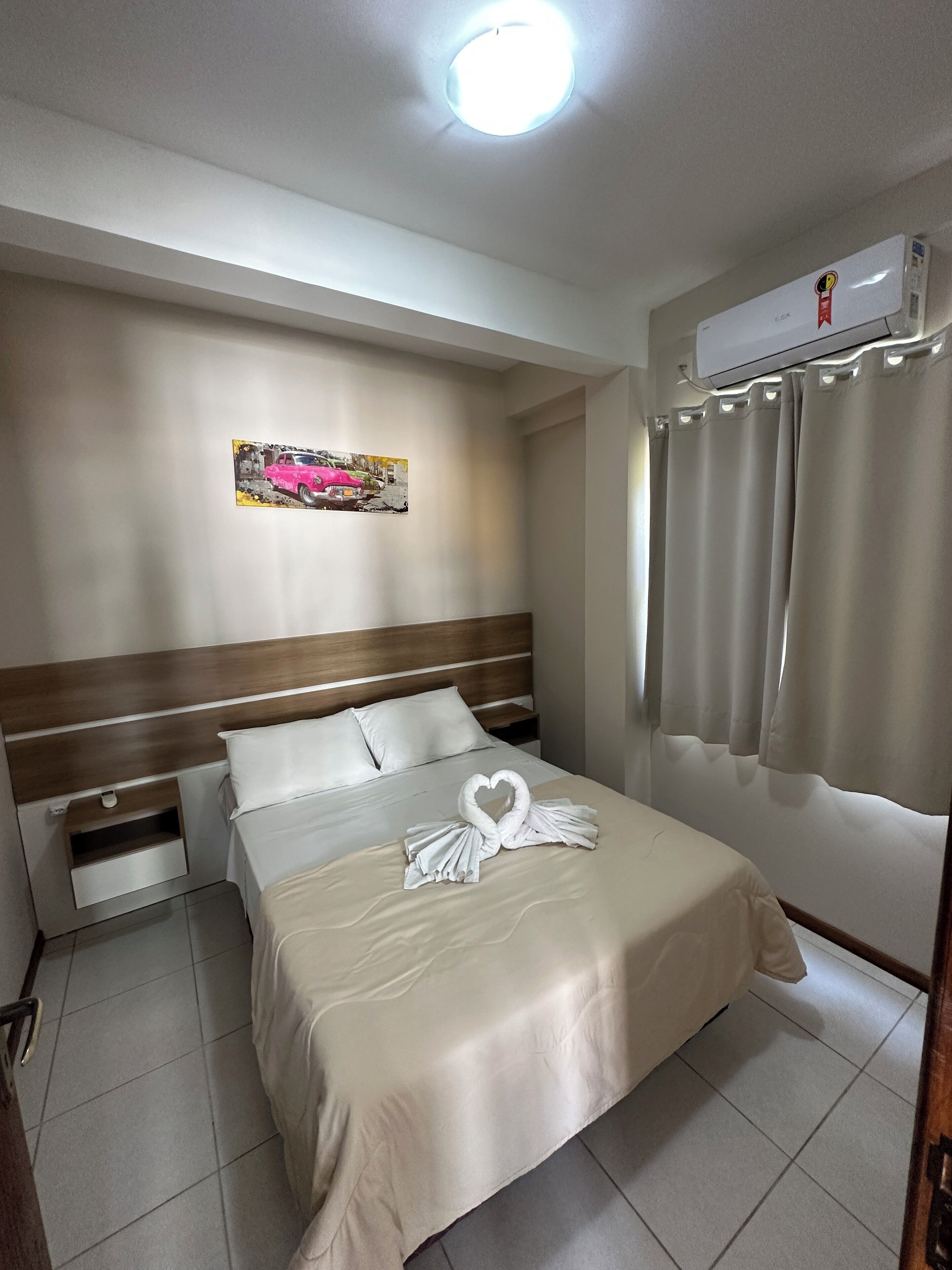 A hotel room with a bed, a nightstand, a wall-mounted air conditioner, and a window with curtains.