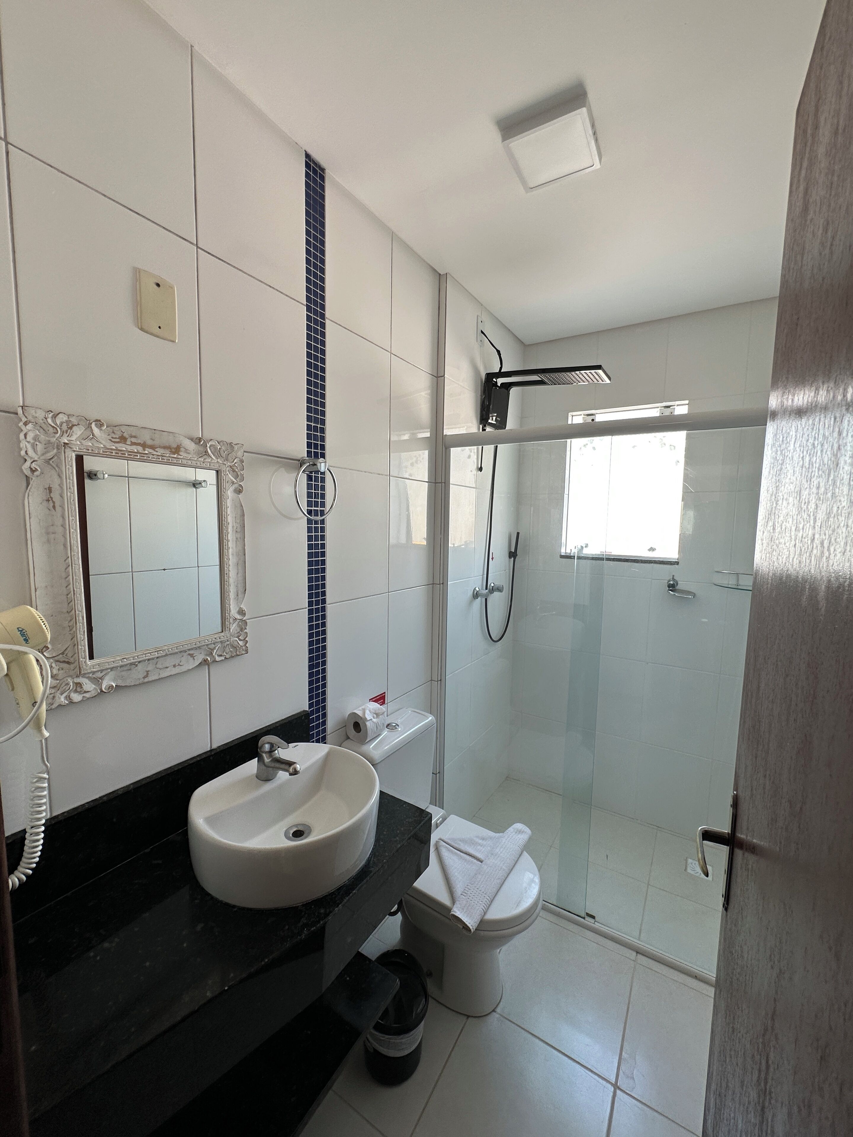 Un baño con lavamanos blanco, un espejo con marco decorativo, toallero, inodoro y zona de ducha con mampara de vidrio.