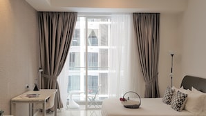 A modern hotel room with a bed, a desk, and a view of the city through large windows.