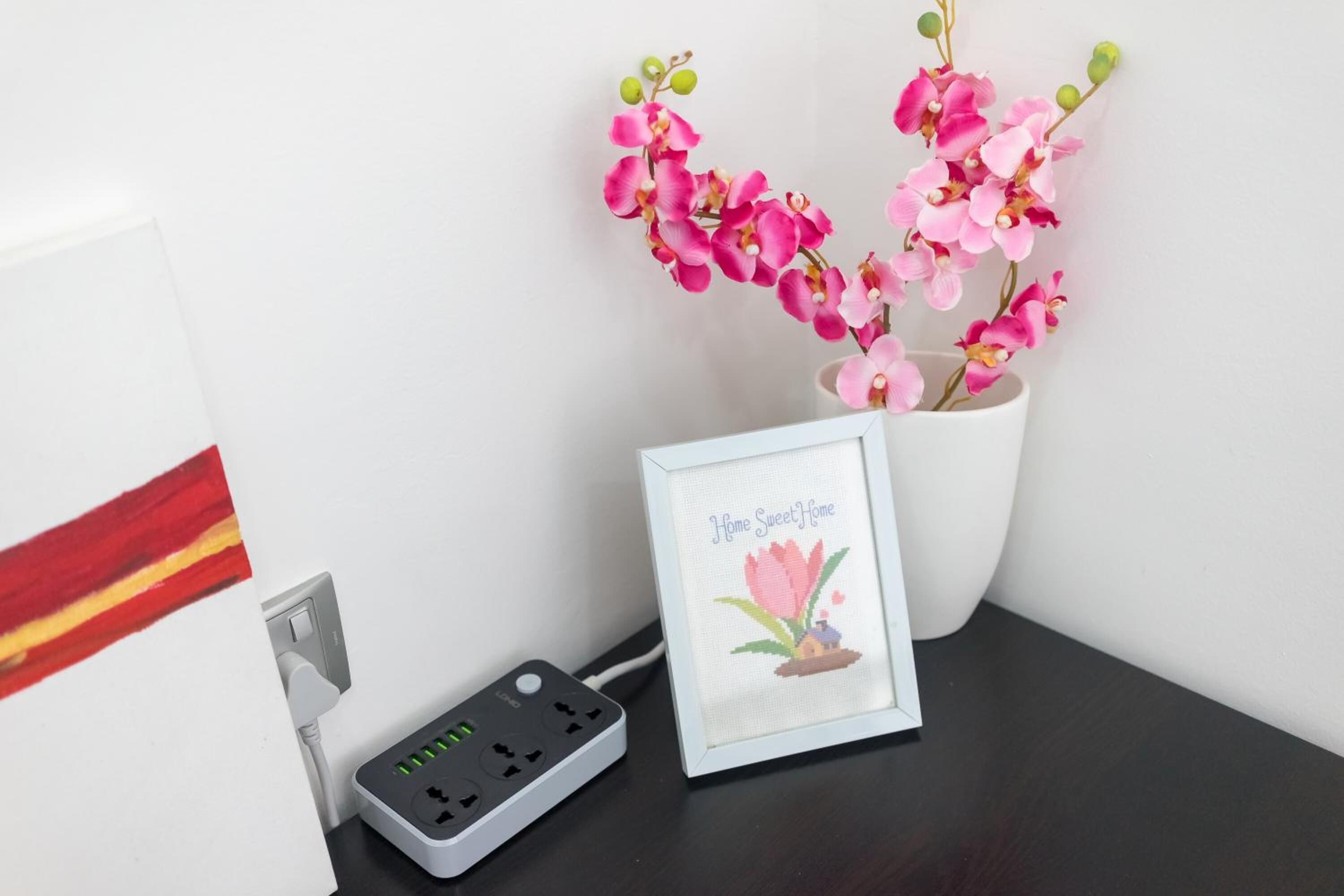 A power strip with multiple outlets, a framed picture with a pink orchid, and a potted plant with pink flowers.