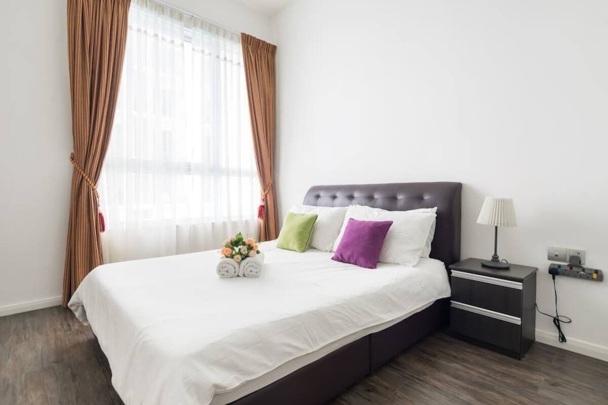 A neatly made bed with white linens, a dark headboard, and decorative pillows. There is a small bedside table with a lamp and a power outlet.