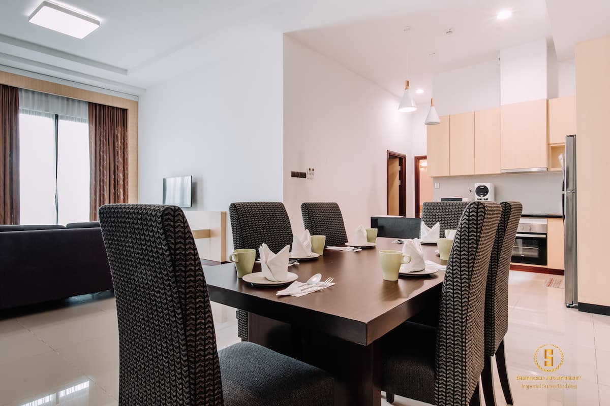 A modern dining area with a rectangular table set for four, surrounded by upholstered chairs. A television is mounted on the wall, and a kitchen is visible in the background.
