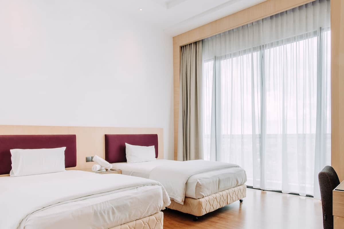 A hotel room with two beds, a wooden headboard, a large window with sheer curtains, and a small table.