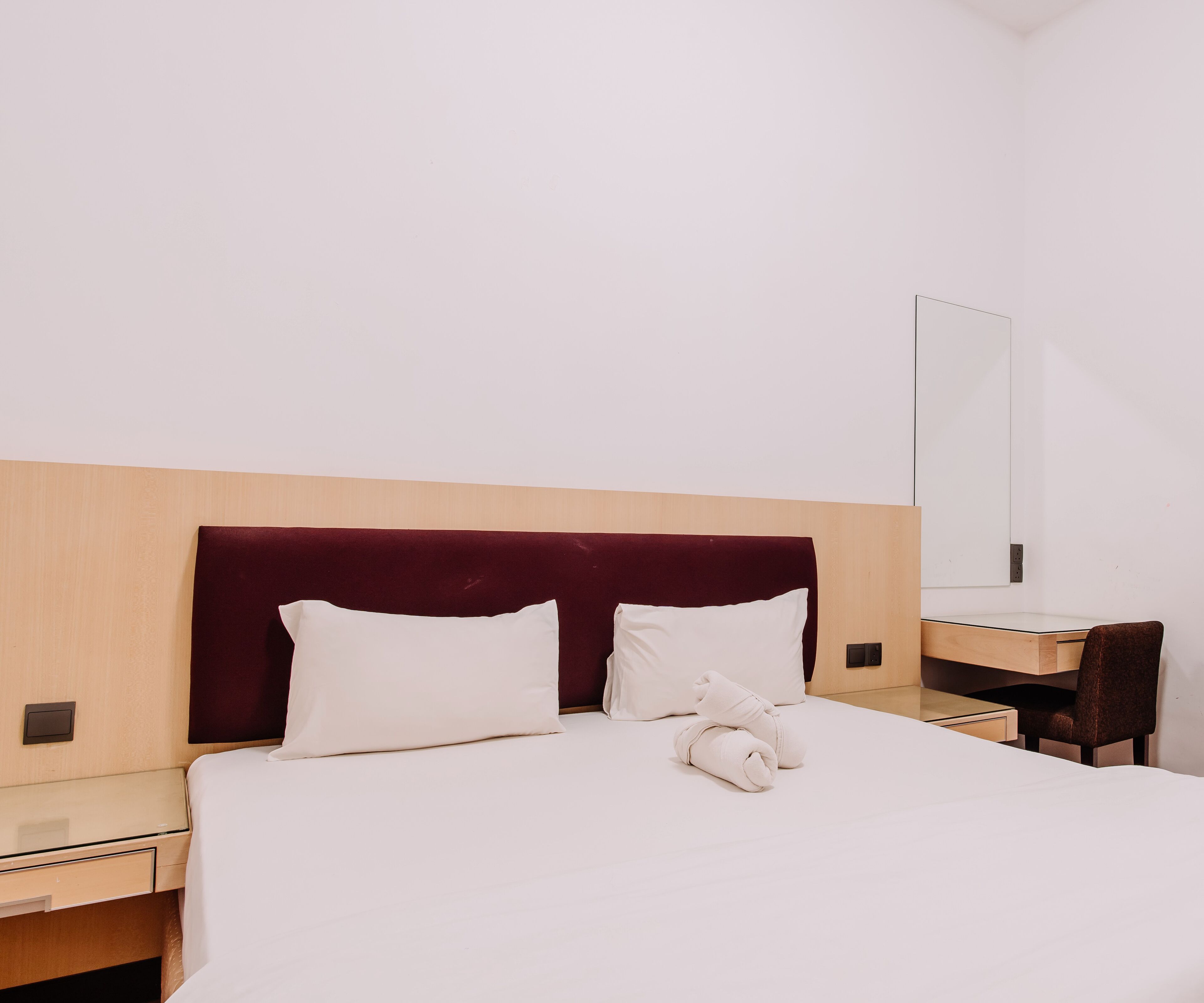 A hotel room with a bed, two pillows, a folded towel, a wooden headboard, a bedside table, and a mirror.