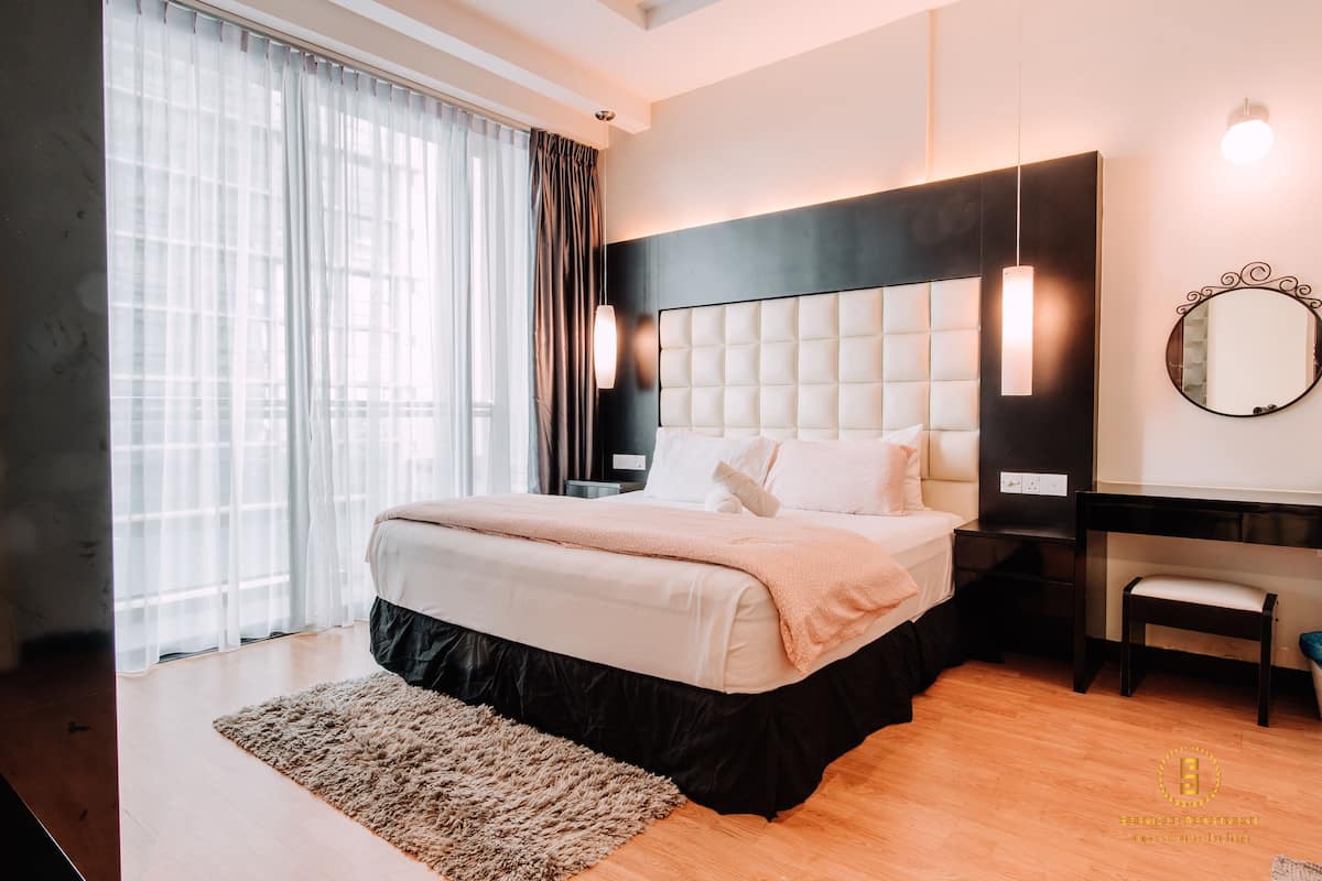 A modern bedroom with a large bed, a desk, and a chair. There is a window with curtains, a wall-mounted mirror, and a light fixture.