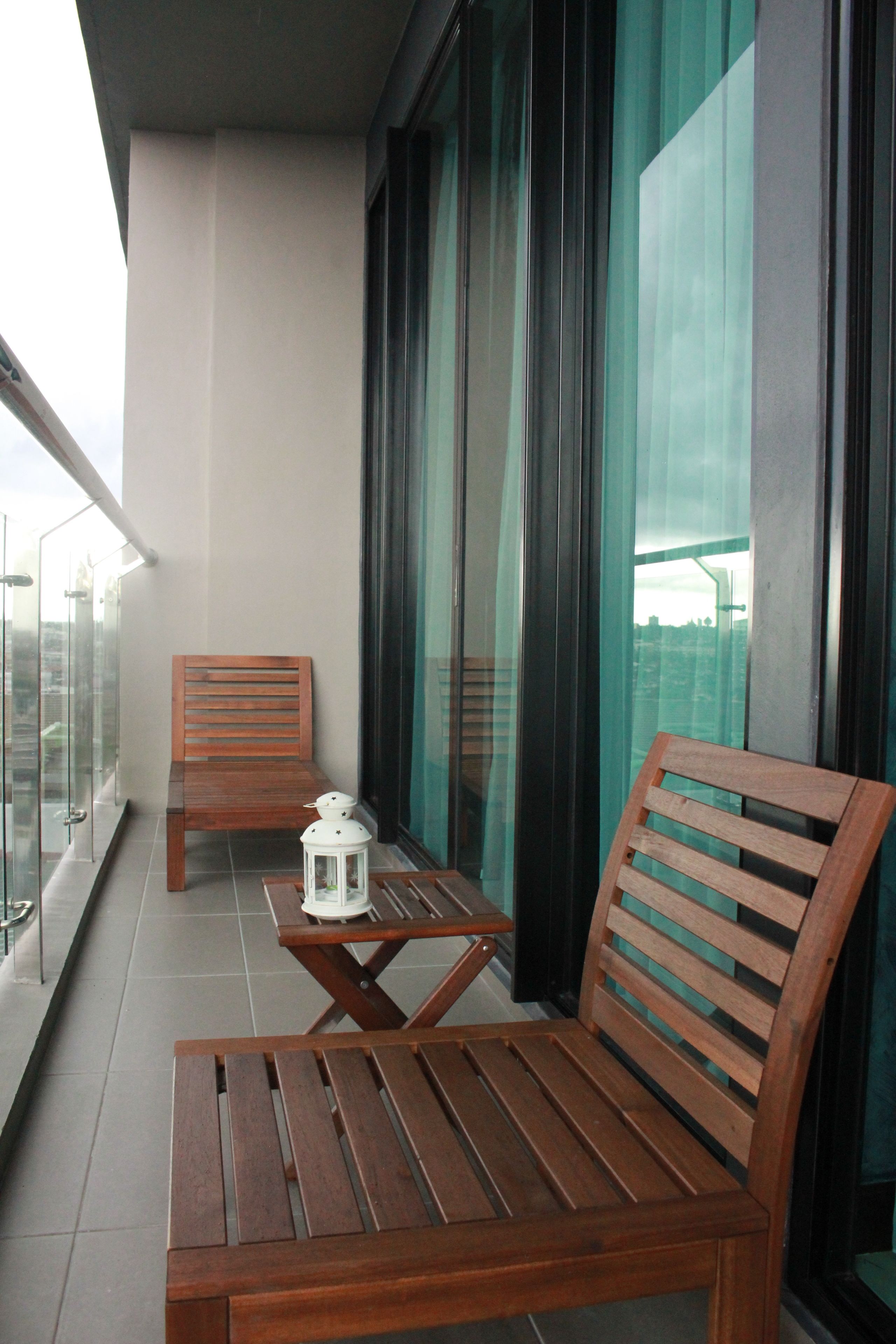 A balcony with a wooden bench and a small table, a white lantern, and a view of the city through large windows.