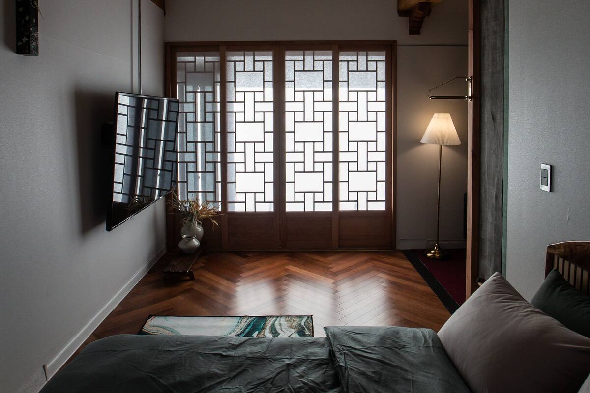 A room with a wooden ceiling, a bed with a green cover, a floor lamp, and a wall-mounted TV.