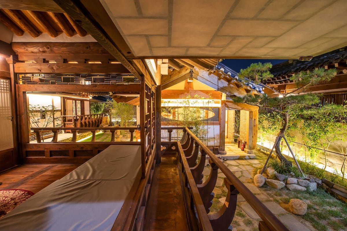 A traditional Japanese-style room with tatami flooring, a low wooden ceiling, and a view of a garden with rocks and plants.