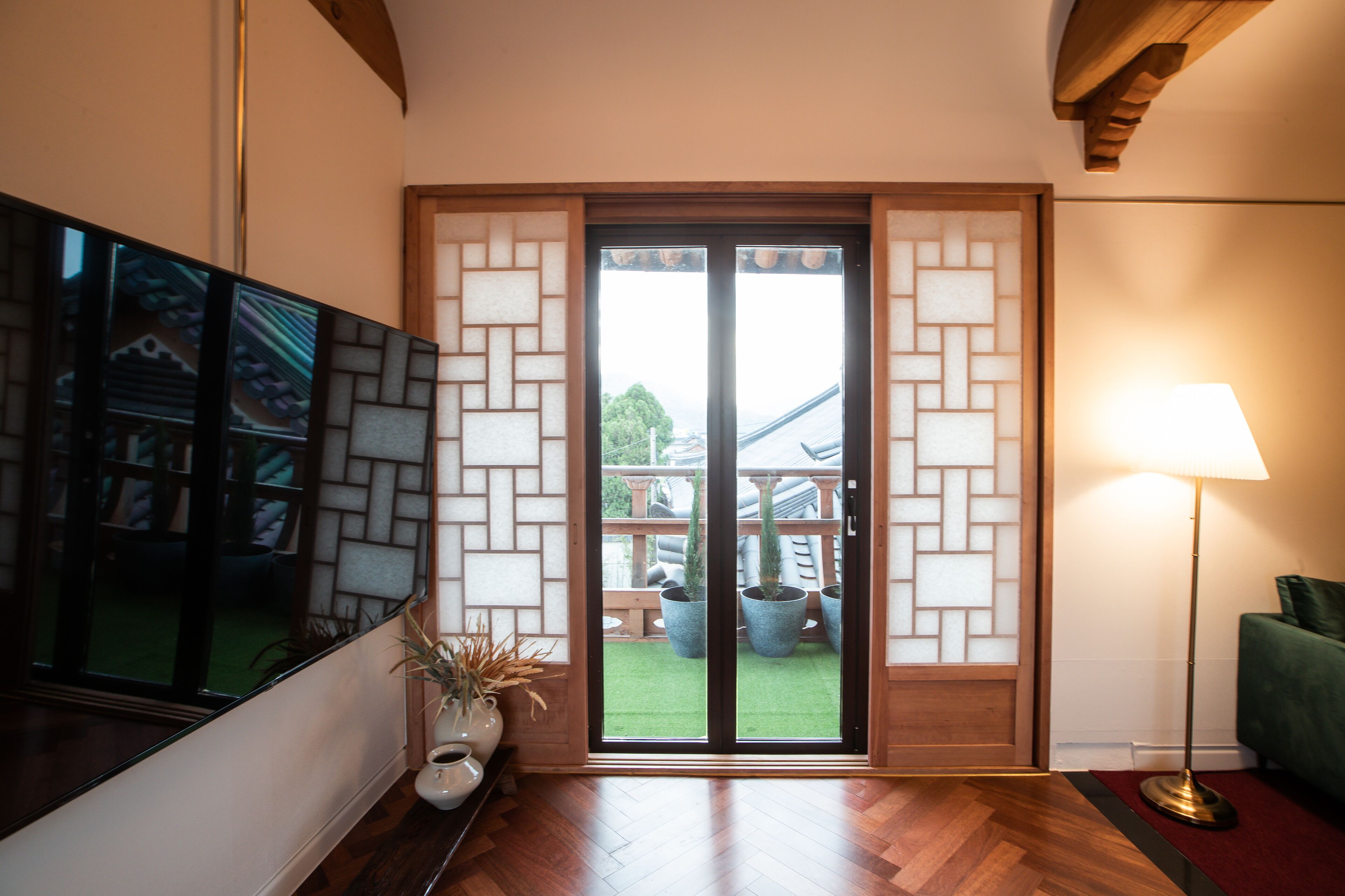 Una stanza con pavimento in legno, una TV fissata al muro, una porta scorrevole in vetro con vista su un giardino e una lampada da terra.