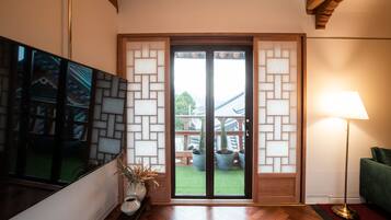 Una stanza con pavimento in legno, una TV fissata al muro, una porta scorrevole in vetro con vista su un giardino e una lampada da terra.