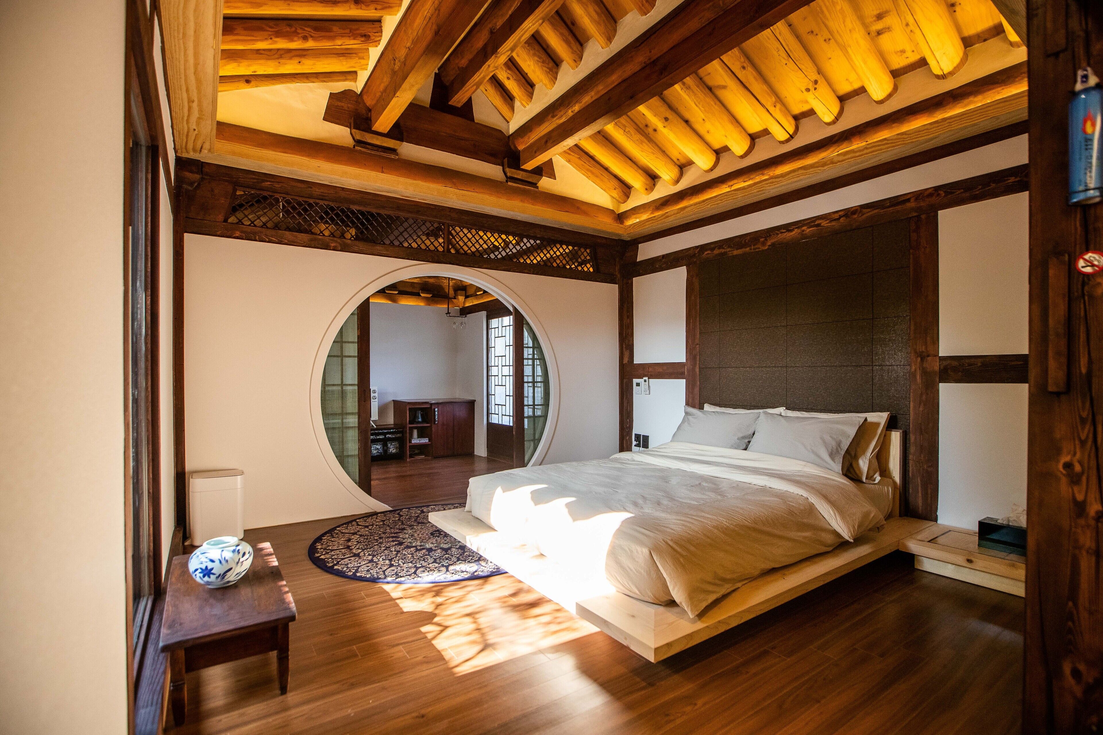 A spacious bedroom with a large bed, wooden beams, a round mirror, and a view of another room.