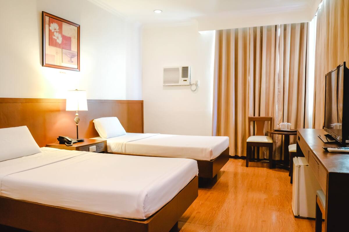 A hotel room with two beds, a desk with a television, a refrigerator, and a window with curtains.