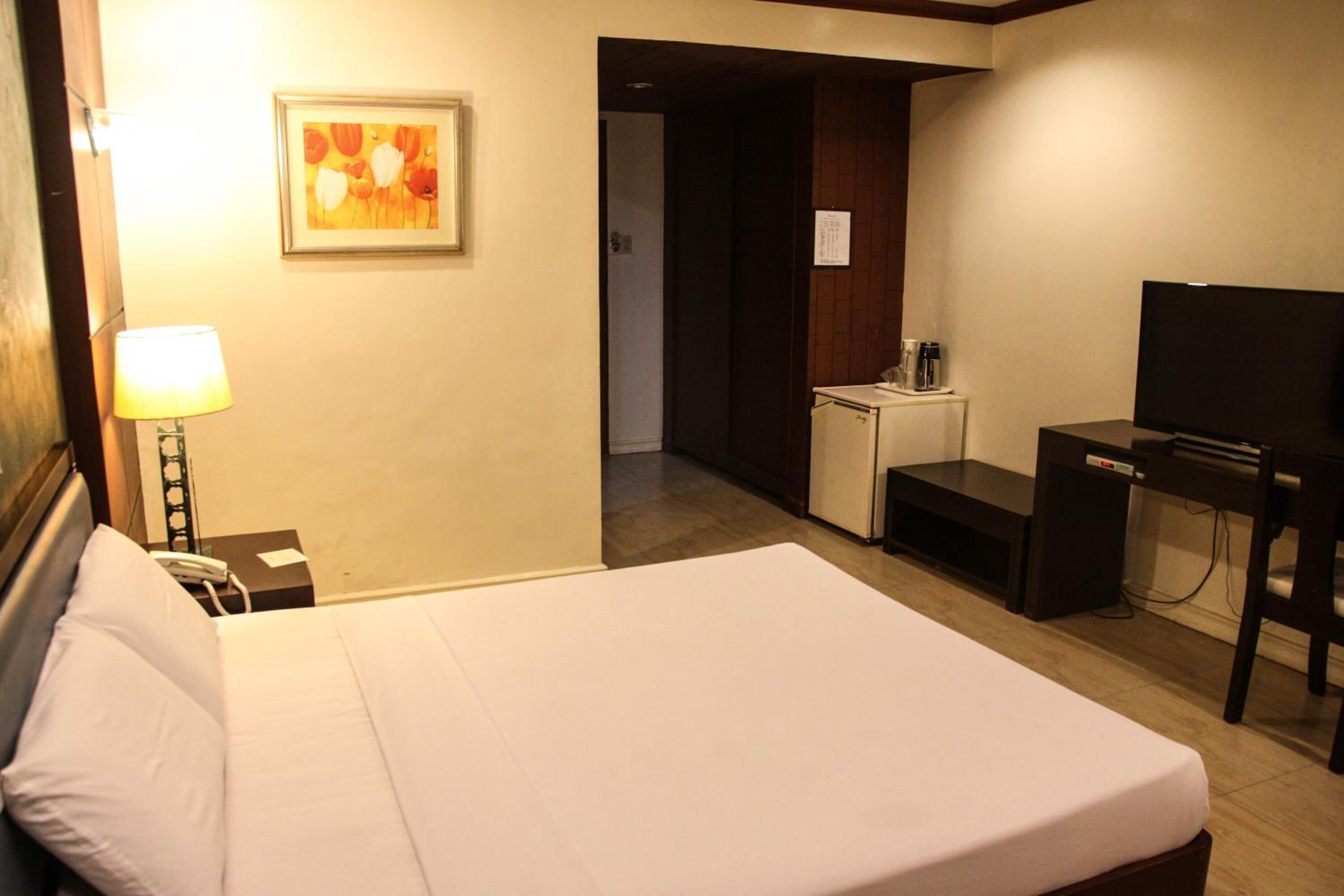 A hotel room with a bed, a desk with a television, a lamp, and a small kitchenette.