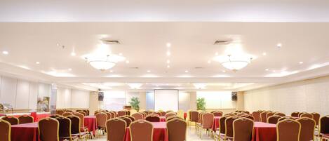A conference room with rows of tables and chairs, equipped with ceiling lights and speakers.