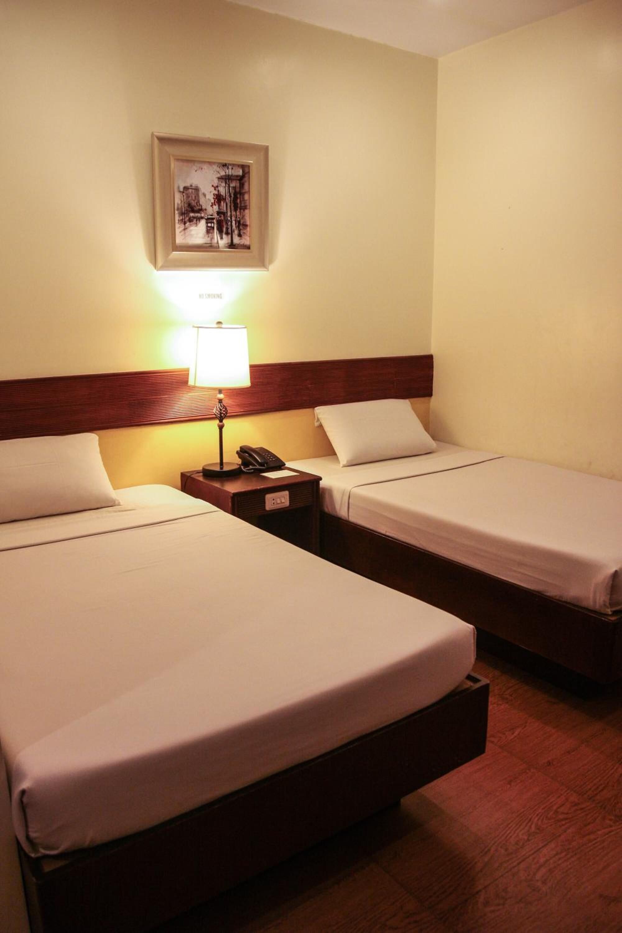A hotel room with two beds, a nightstand with a lamp, a phone, and a framed picture on the wall.