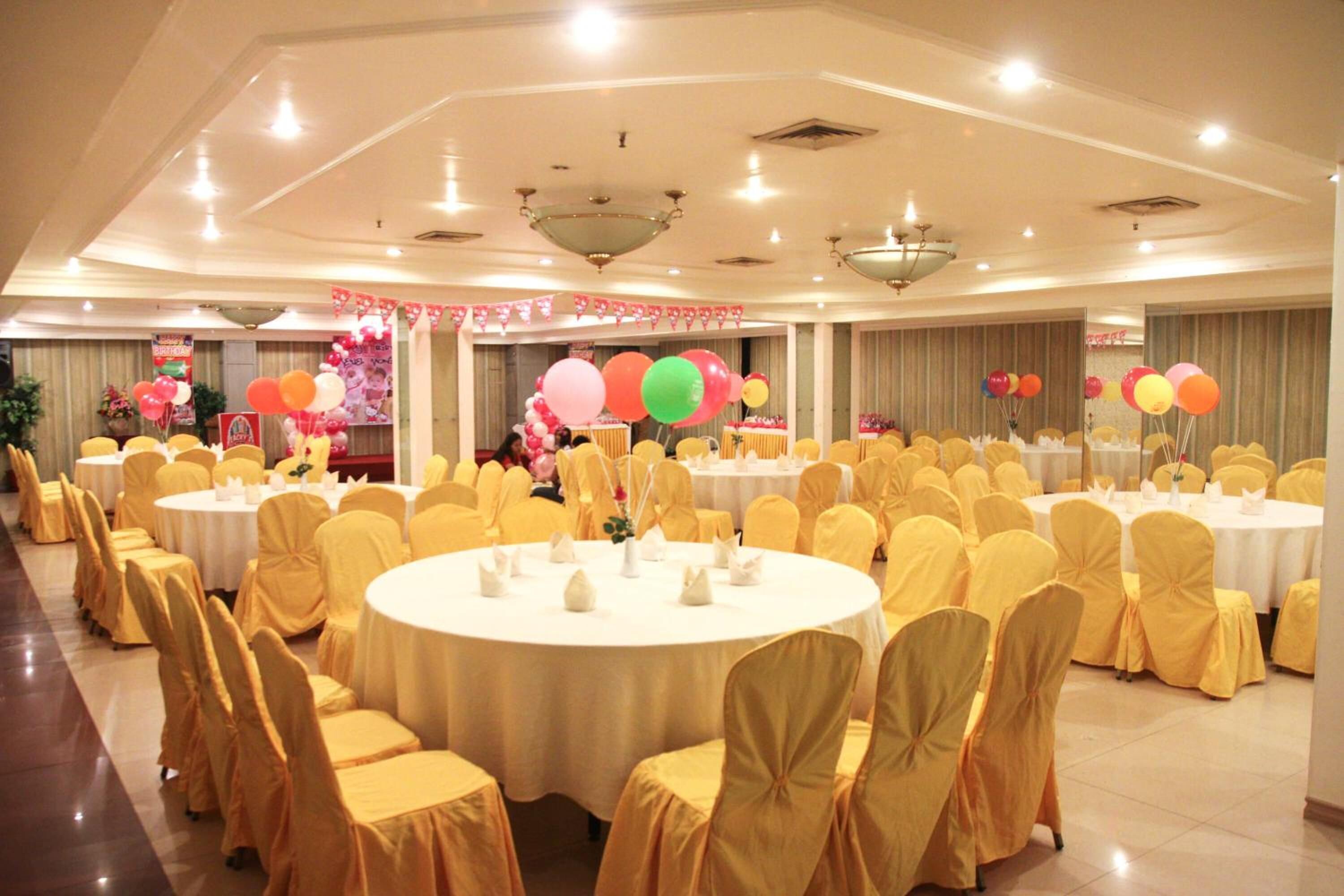 A banquet hall with round tables and yellow-covered chairs, decorated with balloons and floral arrangements.
