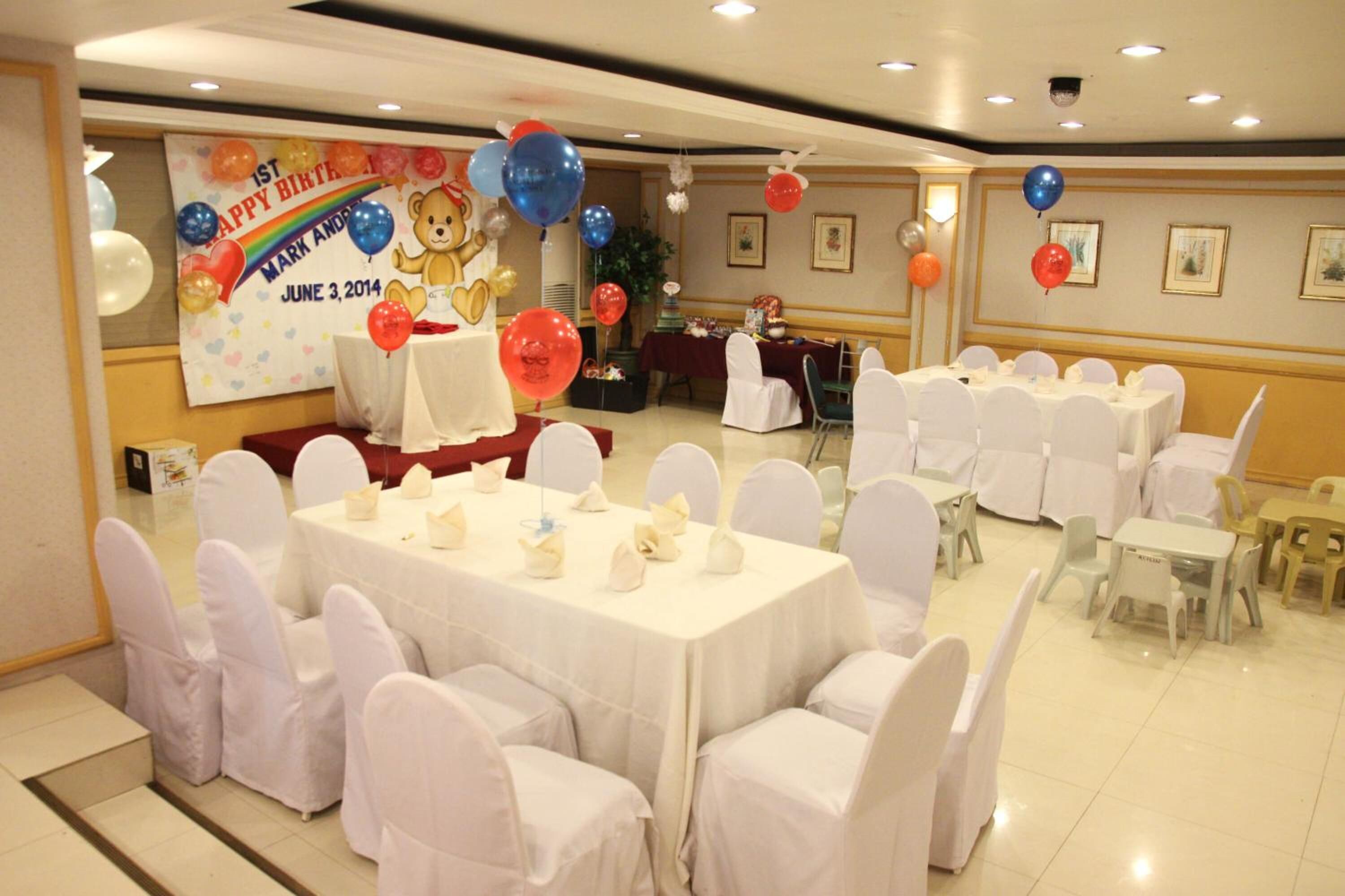 A banquet hall with round tables covered in white tablecloths and white chairs, decorated for a birthday event with balloons and a banner.