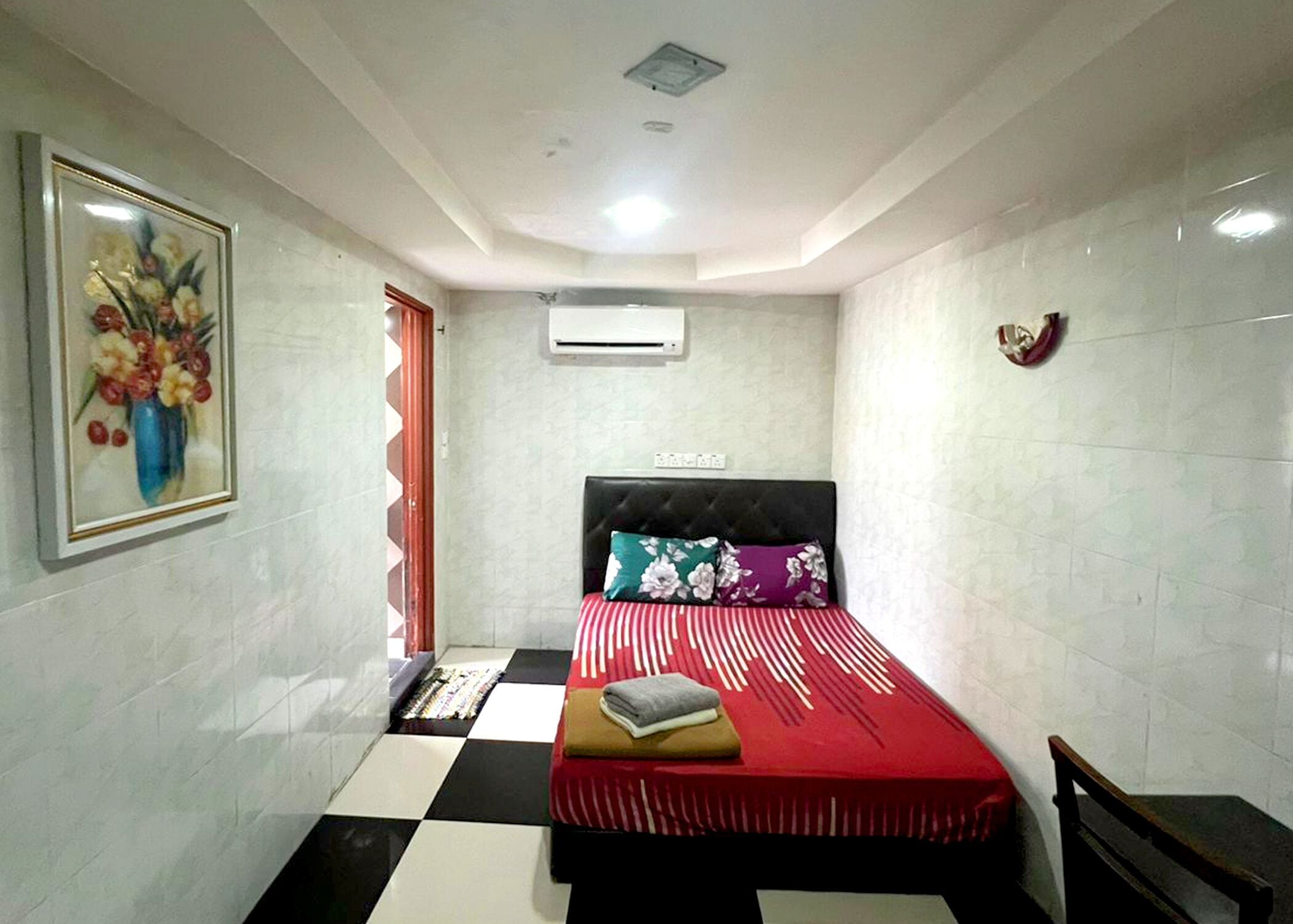 A bedroom with a bed, a framed floral artwork, a red and white striped bedspread, a black headboard, and a white air conditioning unit on the wall.