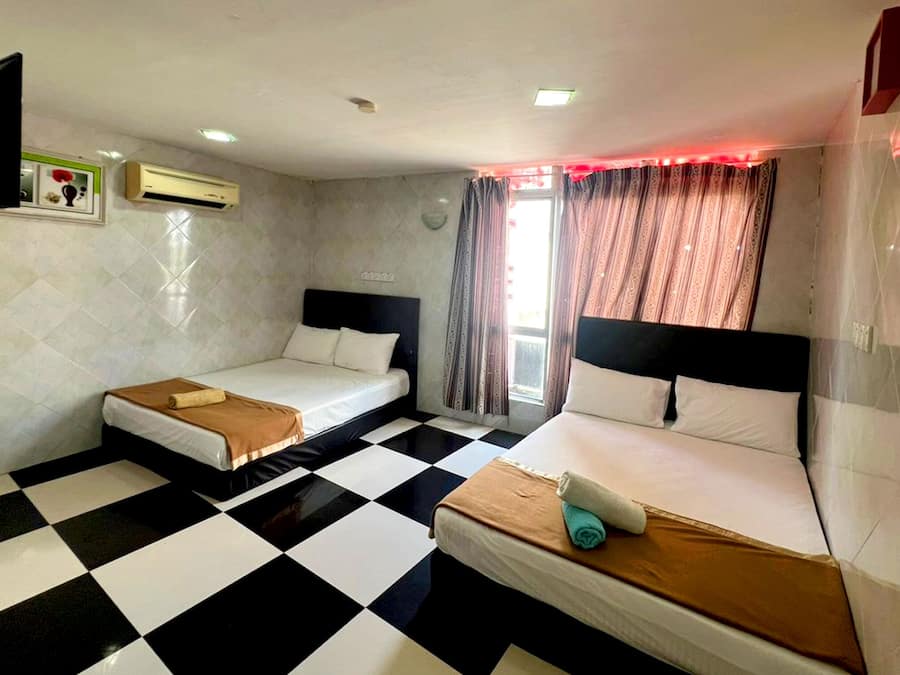 Una habitación de hotel con dos camas individuales, suelo a cuadros, una ventana con cortinas, un equipo de aire acondicionado y un televisor montado en la pared.