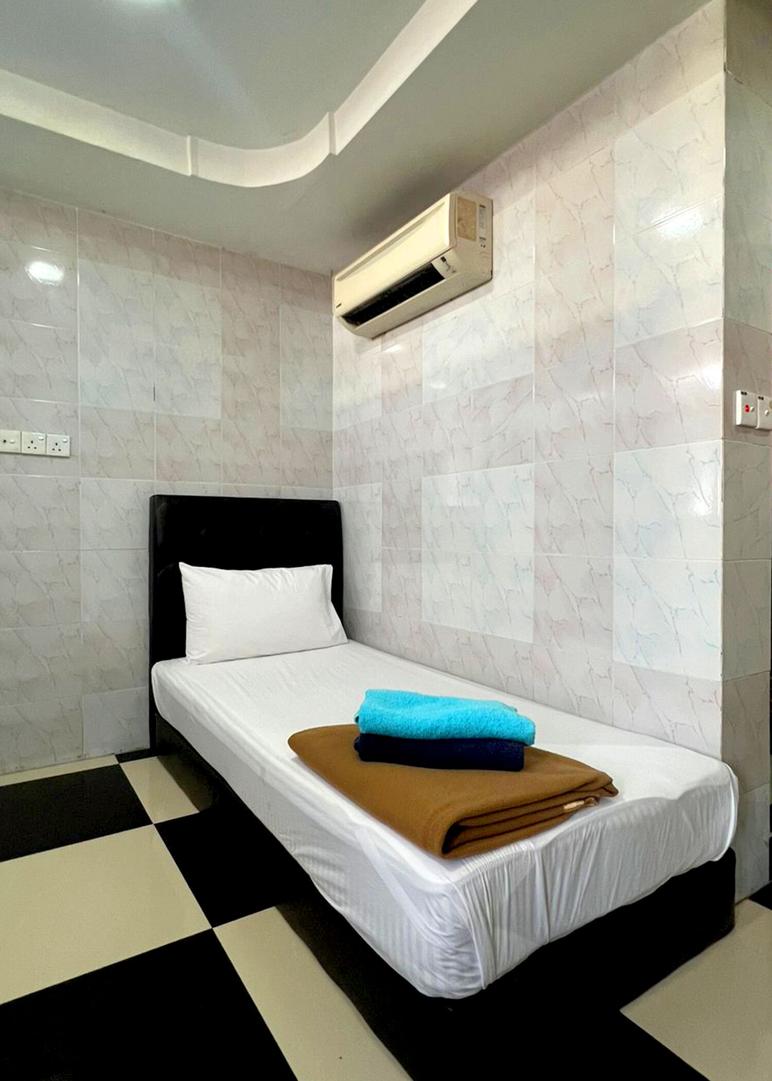 Habitación de hotel con una cama, cabecera negra, colchón blanco, manta marrón y toalla azul.