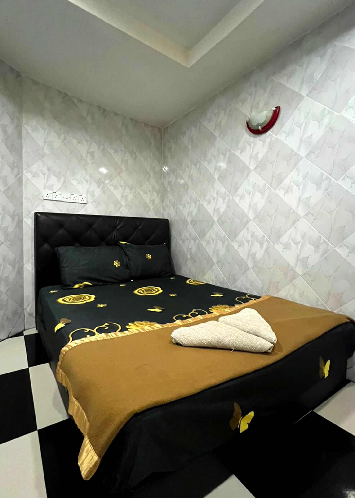 Una cama con cabecera negra y colcha marrón, almohada blanca y un adorno rojo en la pared.