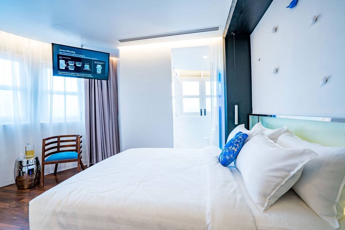 A modern hotel room with a large bed, a desk with a chair, and a screen displaying information.