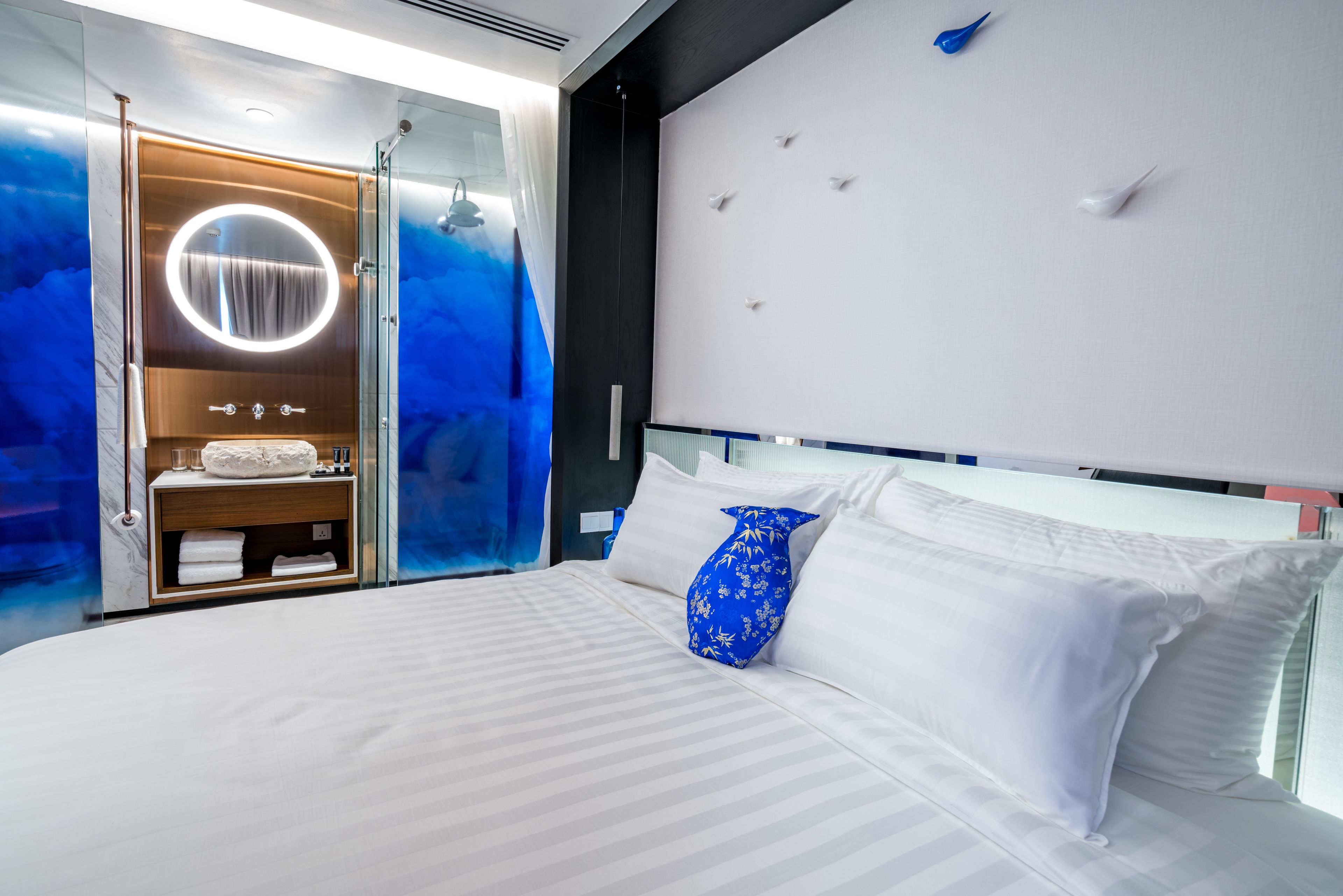 A modern hotel room with a large bed, a glass-enclosed bathroom, and a wall-mounted light fixture.