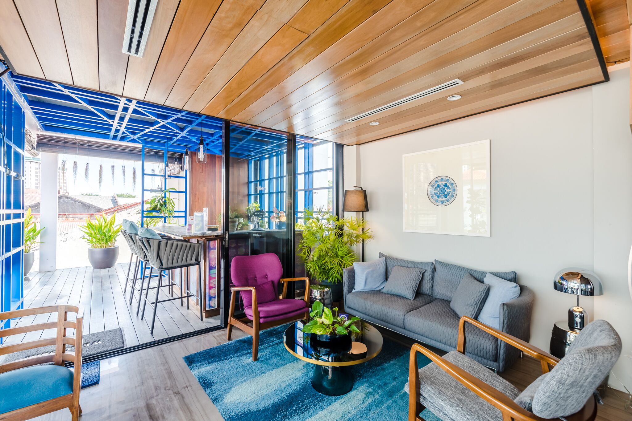 A modern living room with a wooden ceiling, a blue-framed glass door leading outside, a grey sofa, a purple armchair, and a coffee table with plants.