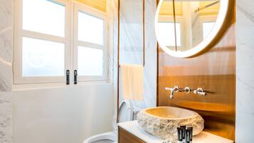 A bathroom with a marble floor, a wooden vanity with a stone sink, a large mirror, and a window with frosted glass.