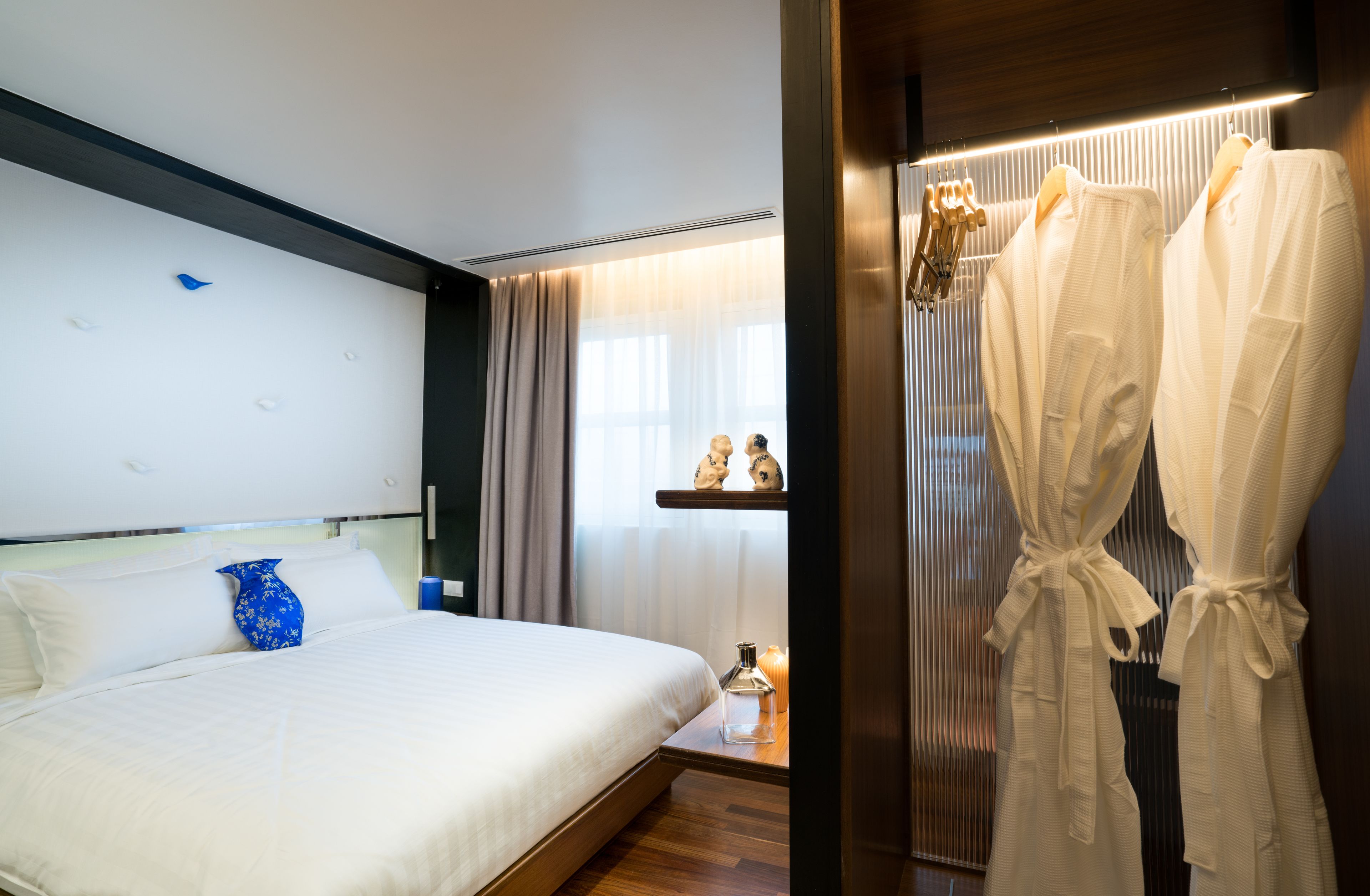 A hotel room with a large bed, a bedside table with a lamp, and a robe rack with robes.