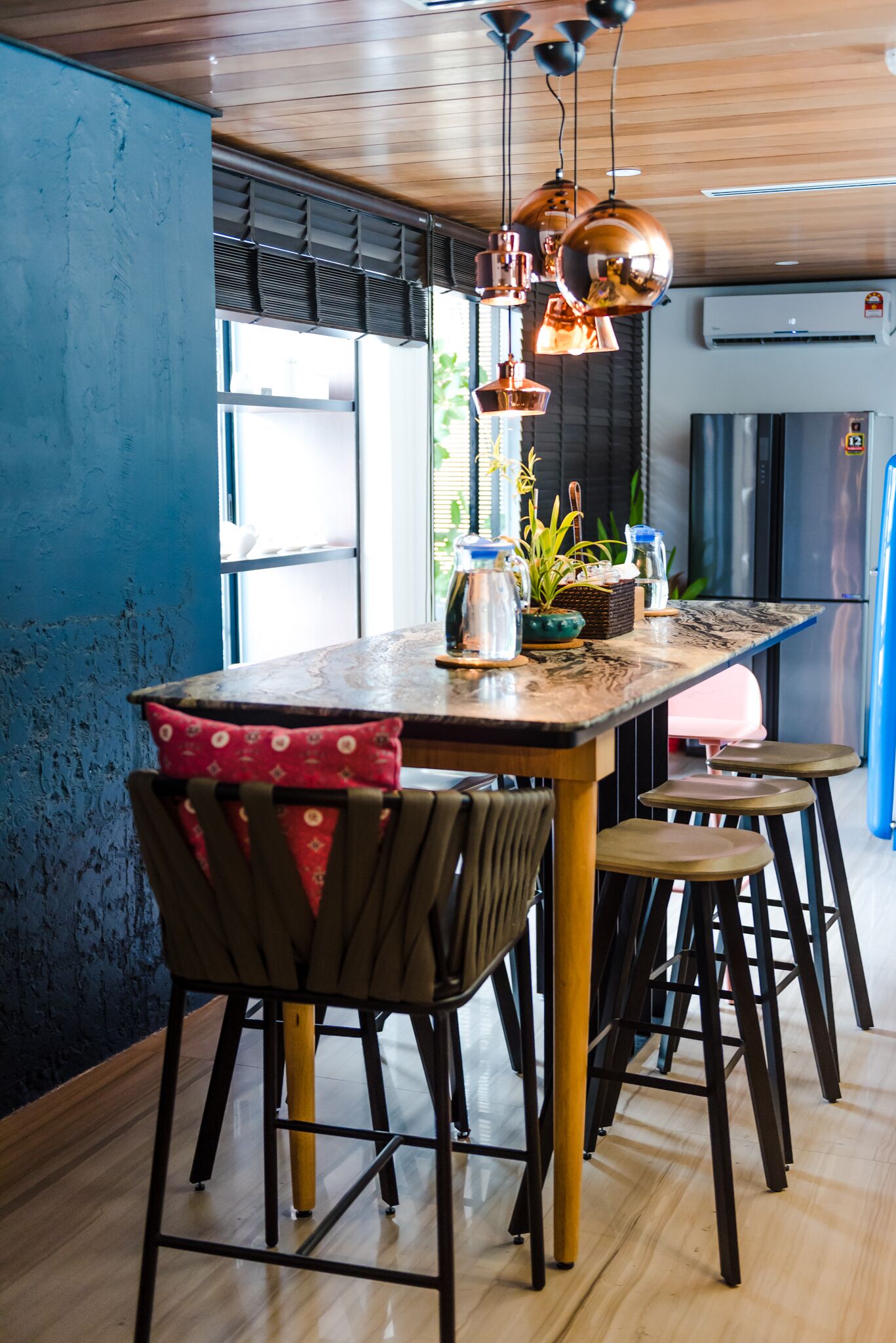 Una cocina moderna con una mesa de madera, taburetes de bar, lámparas colgantes de cobre y vistas al exterior a través de amplios ventanales.