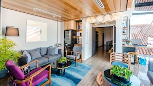 A modern living room with a grey sofa, wooden chairs, a coffee table, and a blue rug. There is a bookshelf and a desk with a laptop in the background. - Tien Hotel (George Town)